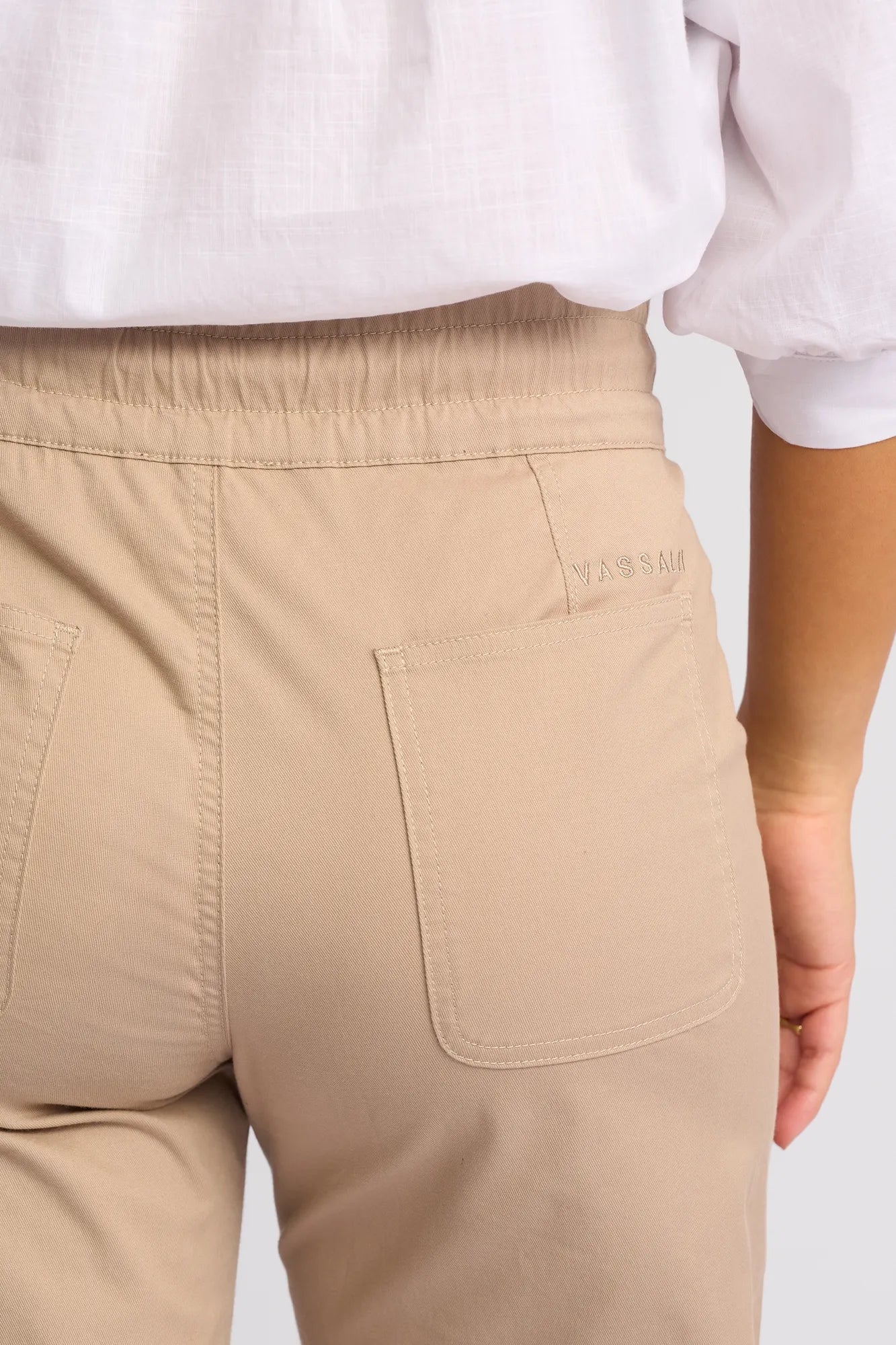 A close-up of the back of a person wearing Vassalli’s Avon Pant in Sand, featuring stretch cotton fabric, an elastic waistband, and two pockets, paired with a white shirt.