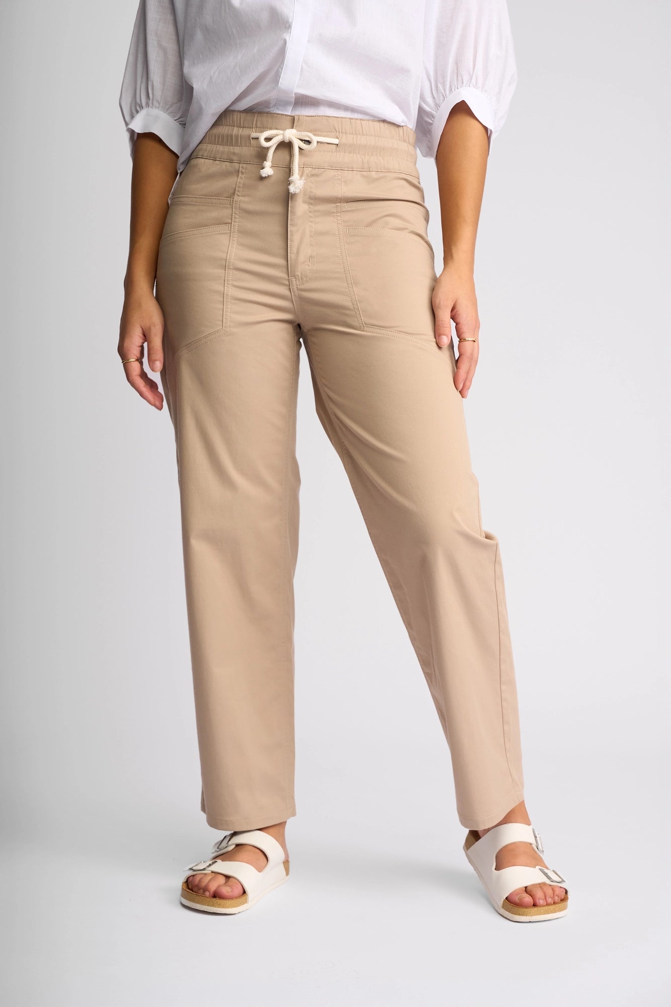 A person wearing Vassalli’s Avon Pant in Sand, featuring straight-leg, ankle-grazer length and utility-inspired styling, pairs them with a white blouse with rolled sleeves and white sandals while standing against a plain light background.