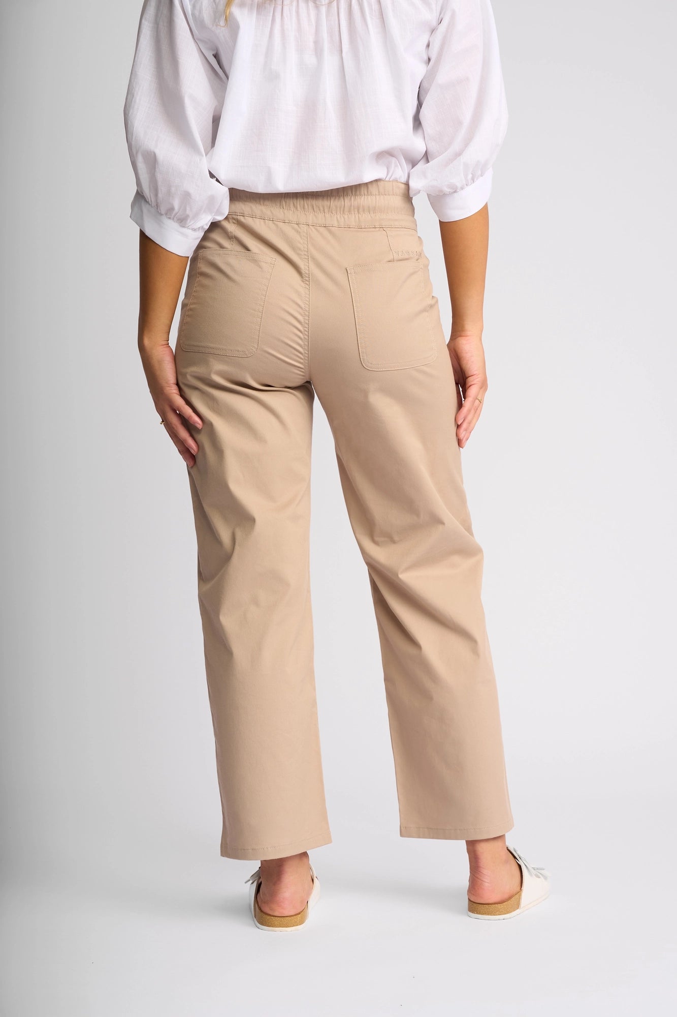 A person wearing the Vassalli Avon Pant in Sand—beige straight-leg ankle-grazer pants crafted from stretch cotton with subtle utility-inspired styling for a modern yet effortless look—and a white blouse, shown from the back against a plain light background.