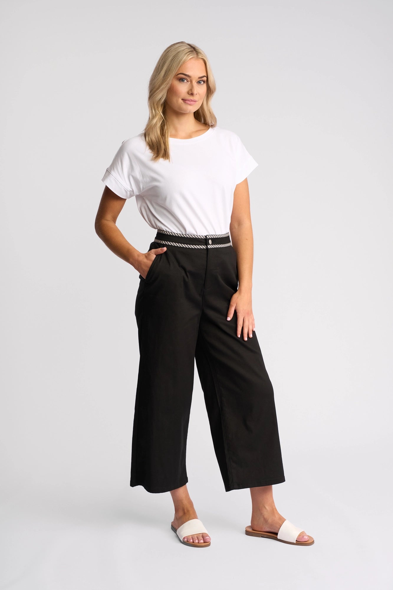 A woman stands against a plain background, wearing the Vassalli Atlanta Jean in Black Denim with a white t-shirt and a braided belt. She has on white slide sandals, her blonde hair is loose, and she stands with her hands in her pockets, smiling softly.