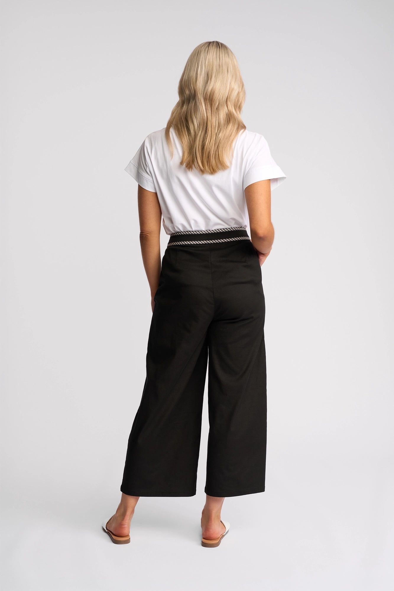 A woman with long blonde hair, wearing a white short-sleeve shirt, Vassalli’s Atlanta Jean in Black Denim—featuring wide-leg stretch denim and a belt—and sandals, stands facing away in a studio setting.