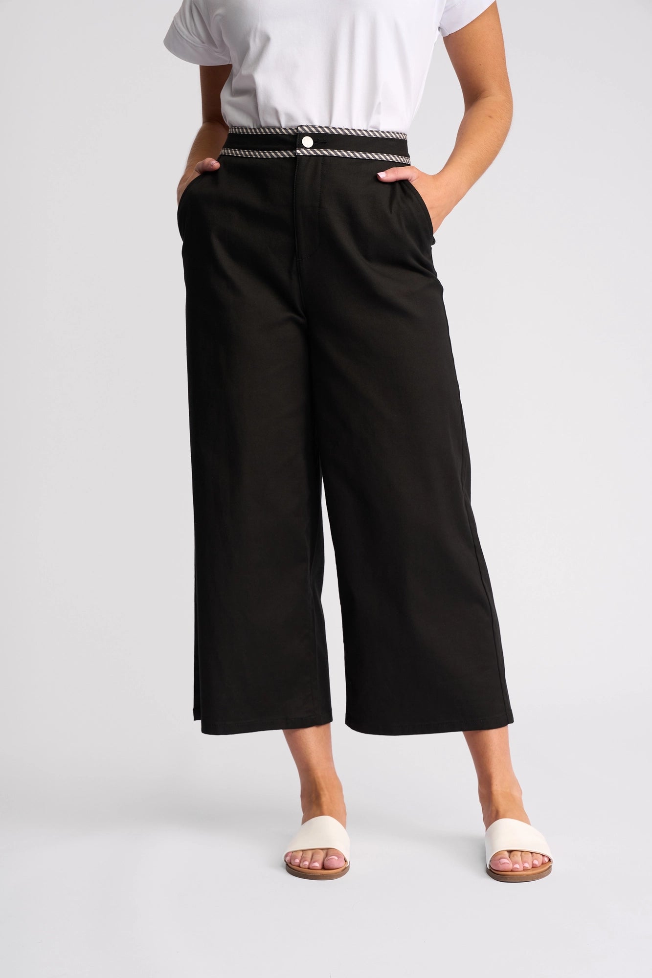 Wearing a white shirt and Vassalli’s Atlanta Jean - Black Denim, which features a wide-leg cropped fit and striped waistband, paired with white slide sandals, the person stands with hands in pockets against a plain background.