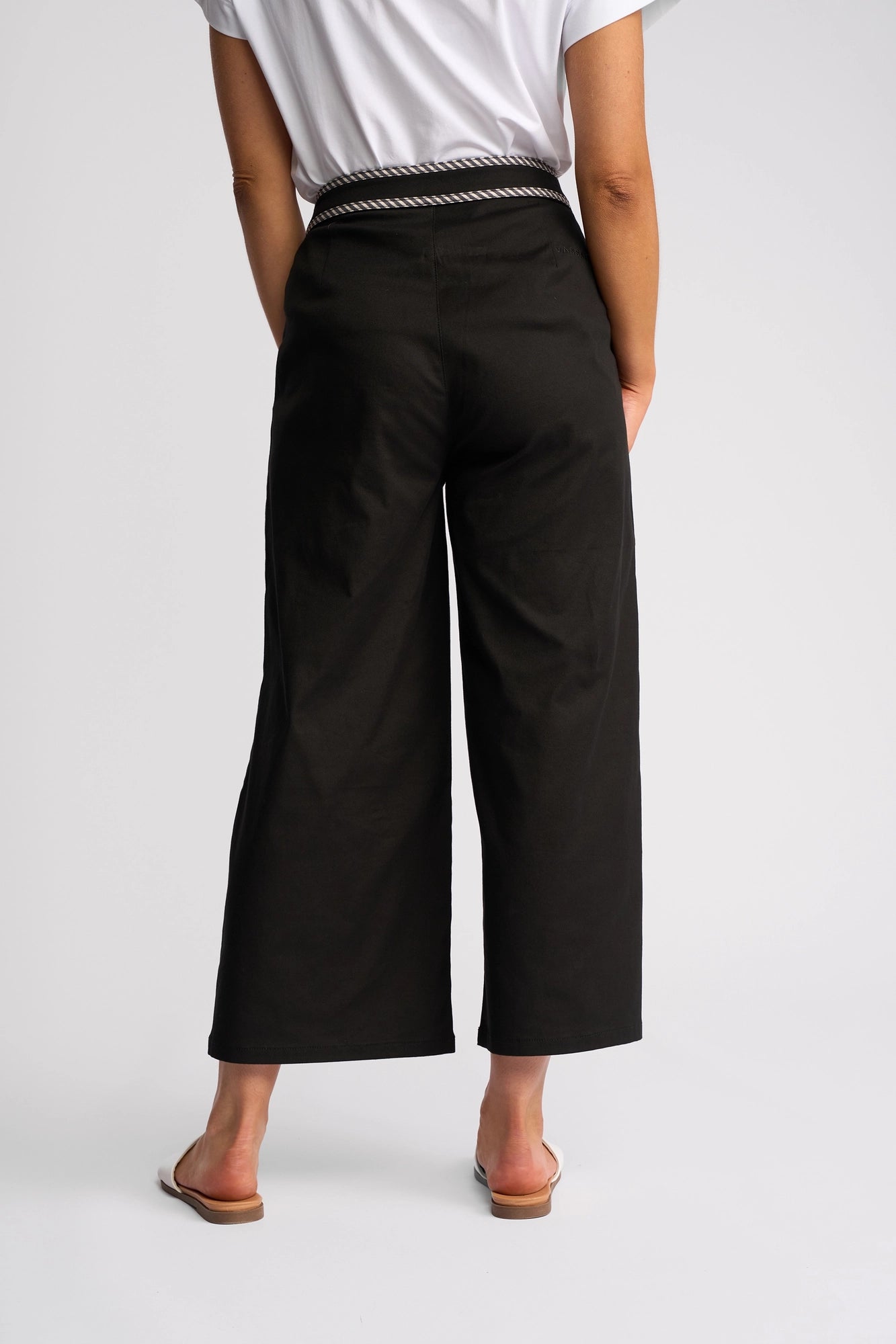 A person stands facing away, wearing a white shirt, white sandals, and the Vassalli Atlanta Jean in black denim, which features stretch fabric, a wide-leg silhouette with a slightly cropped length, and a striped waistband.