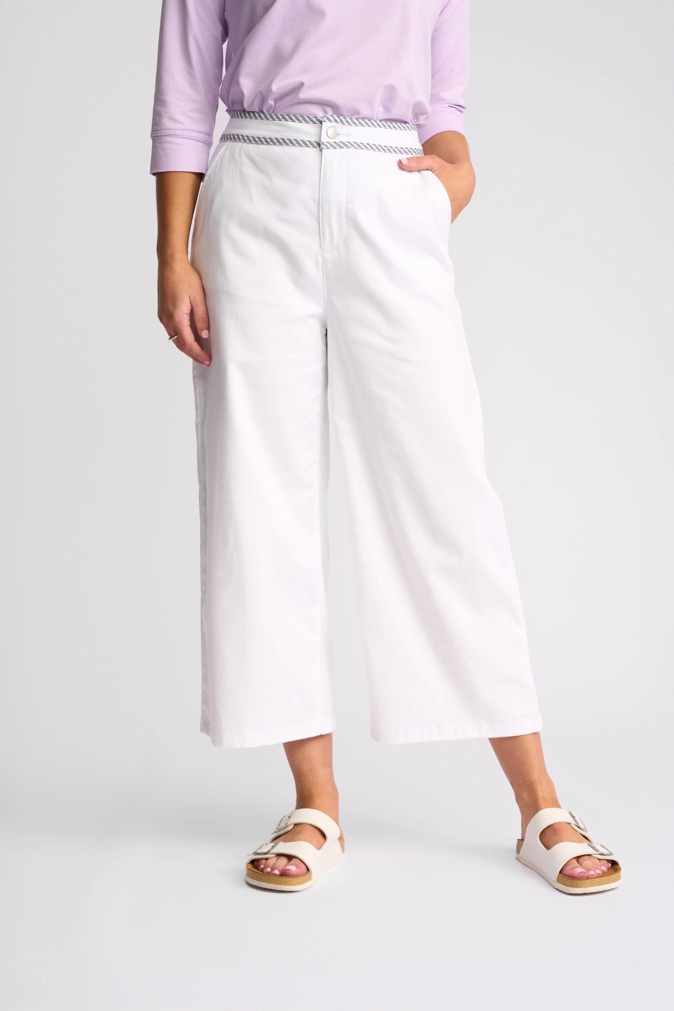 Wearing the Vassalli Atlanta Jean in White Denim, styled with a light purple long-sleeve shirt, a patterned belt, and white sandals. The photo is cropped at the shoulders, with one hand placed in a pocket.