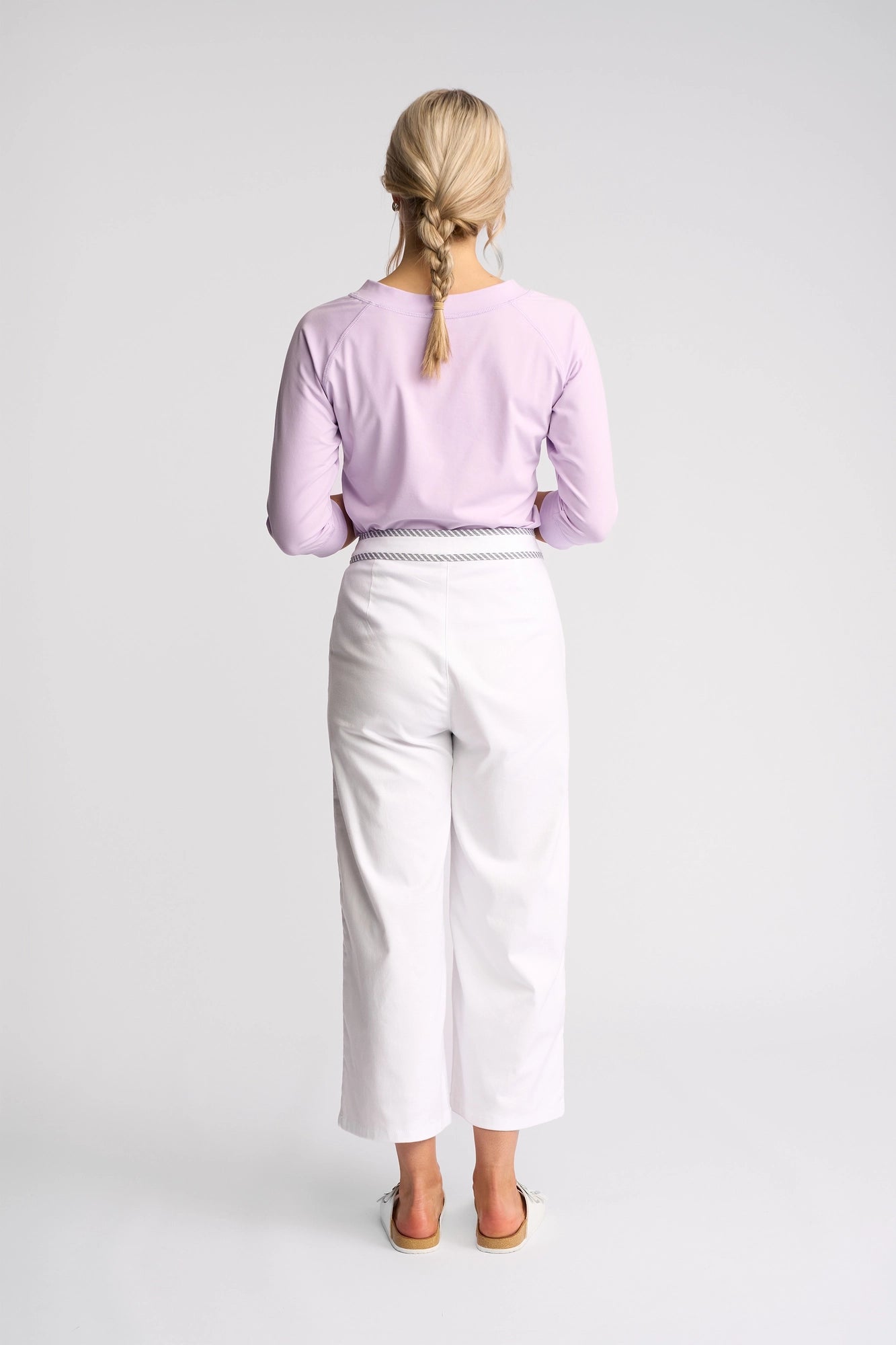 A woman with braided blonde hair stands with her back to the camera against a plain white background, wearing Vassalli’s Atlanta Jean in White Denim styled with a striped waistband, paired with a light purple long-sleeve shirt and sandals.