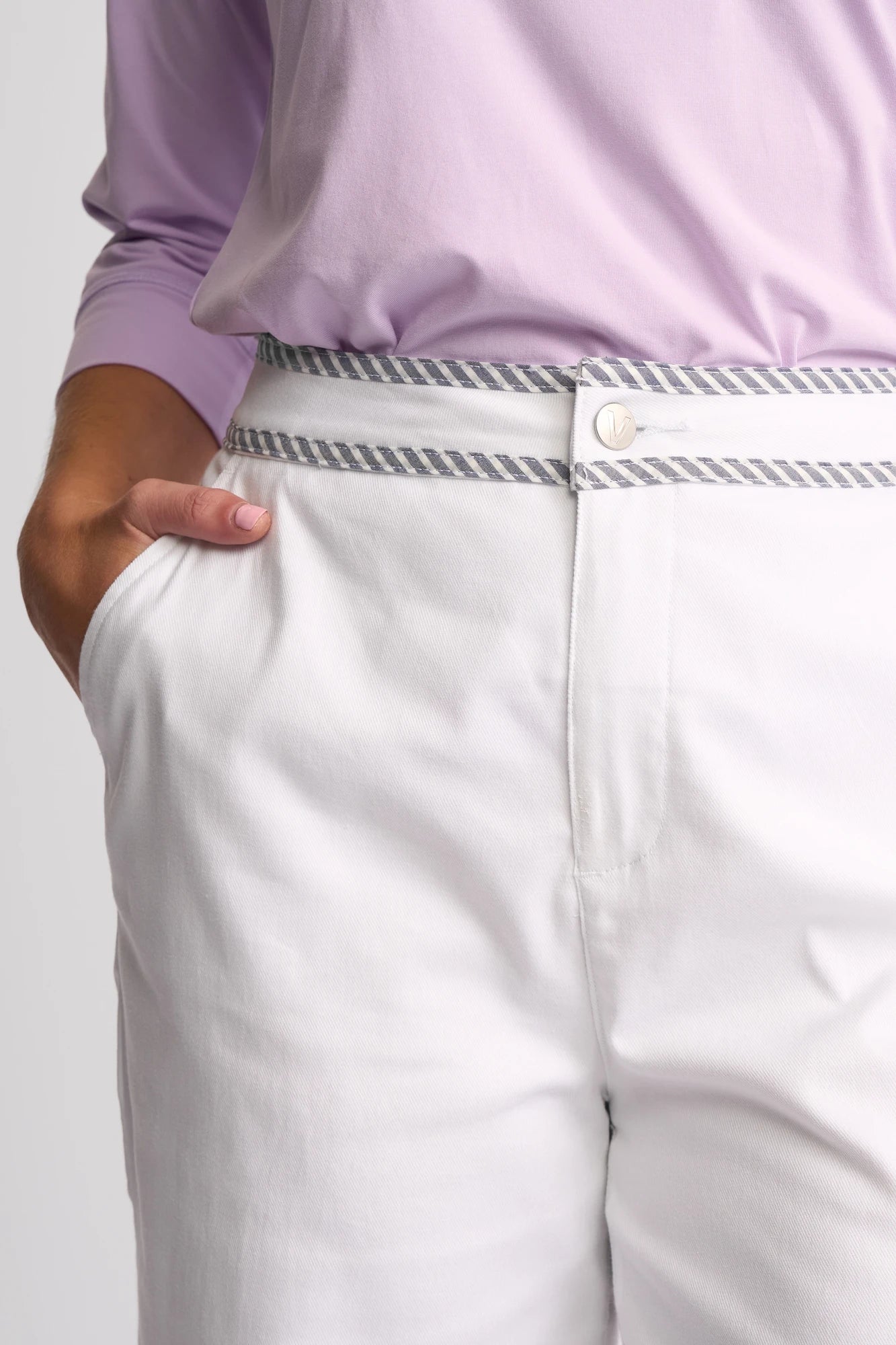 Wearing the Vassalli Atlanta Jean in White Denim, a person stands with one hand in their pocket, pairing the jeans—which feature a striped waistband—with a light purple long-sleeve top.
