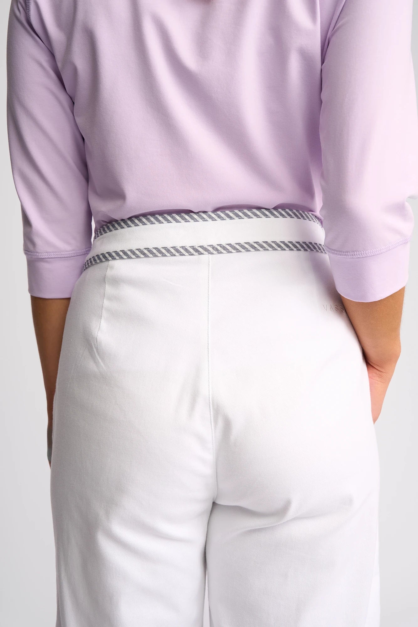 A person shown from the back is wearing Vassalli’s Atlanta Jean in White Denim, featuring a gray and white striped waistband, paired with a light purple top.