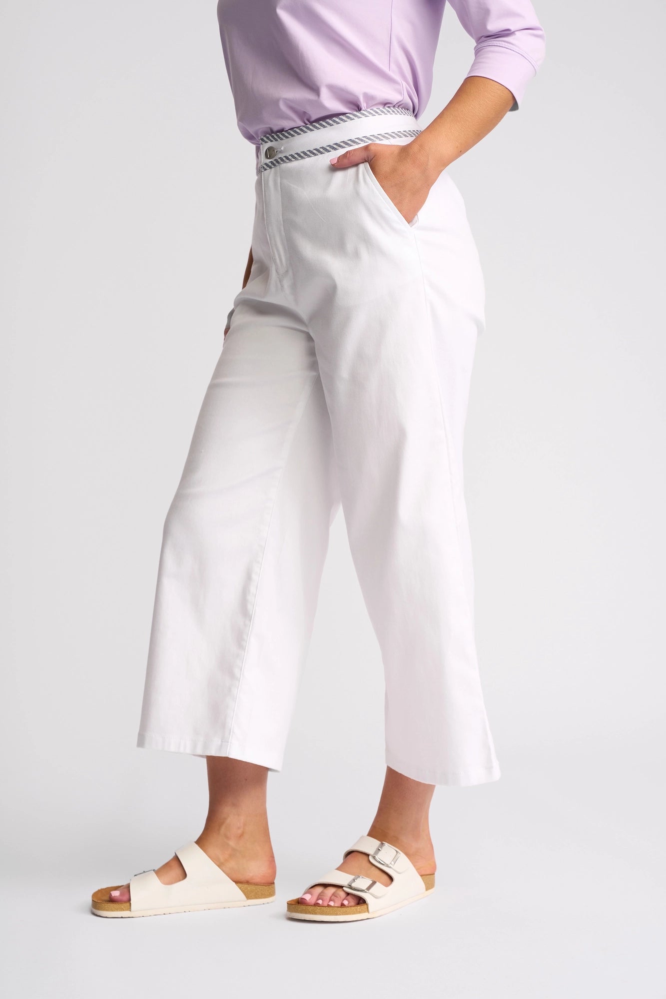 A person stands against a plain background with one hand in their pocket, wearing Vassalli’s Atlanta Jean in White Denim paired with a light purple top and beige sandals.