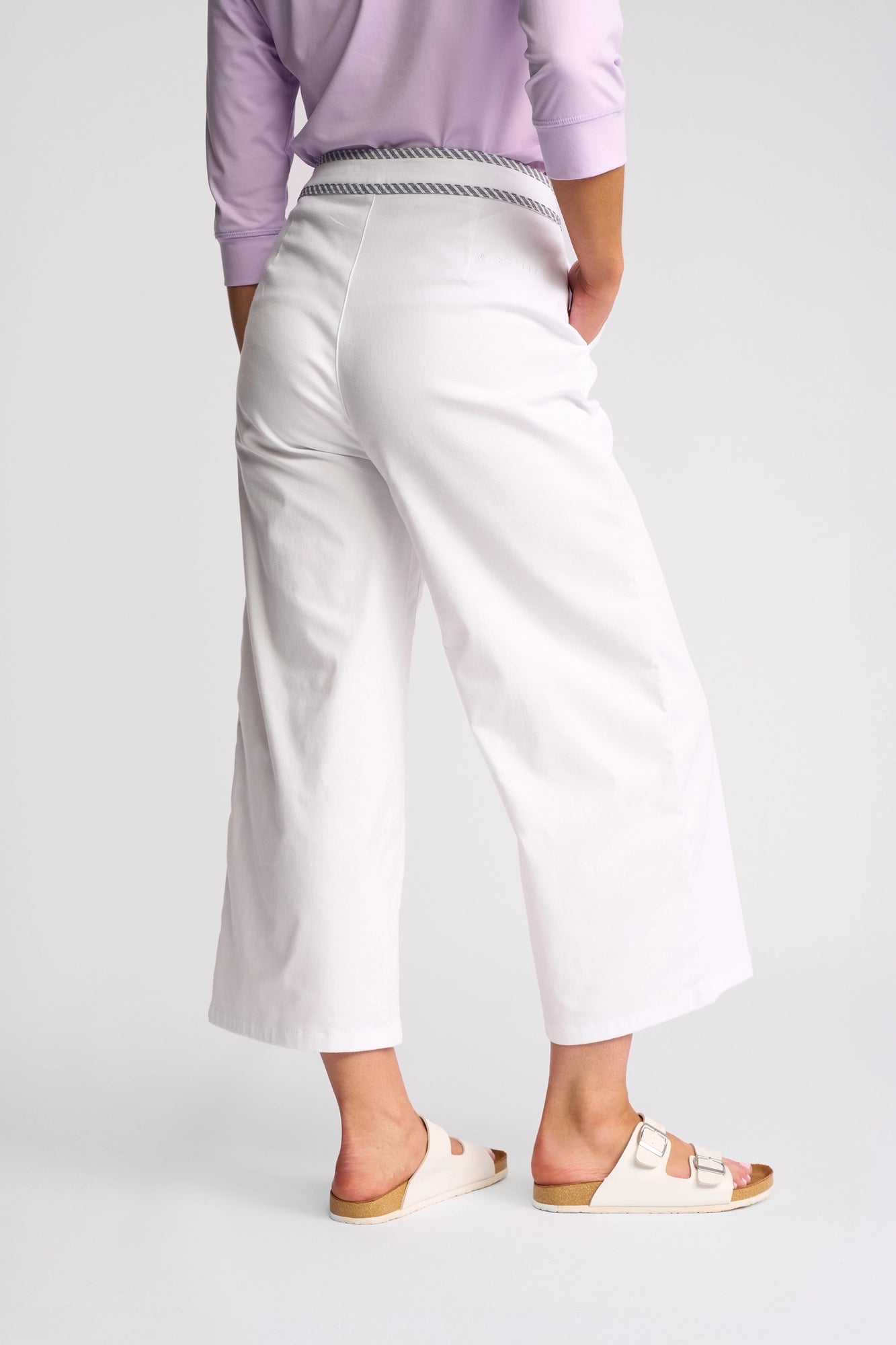 A person stands with their back to the camera against a plain light background, wearing a lavender long-sleeve top, the Vassalli Atlanta Jean in White Denim featuring a subtle striped waistband, and beige two-strap sandals.