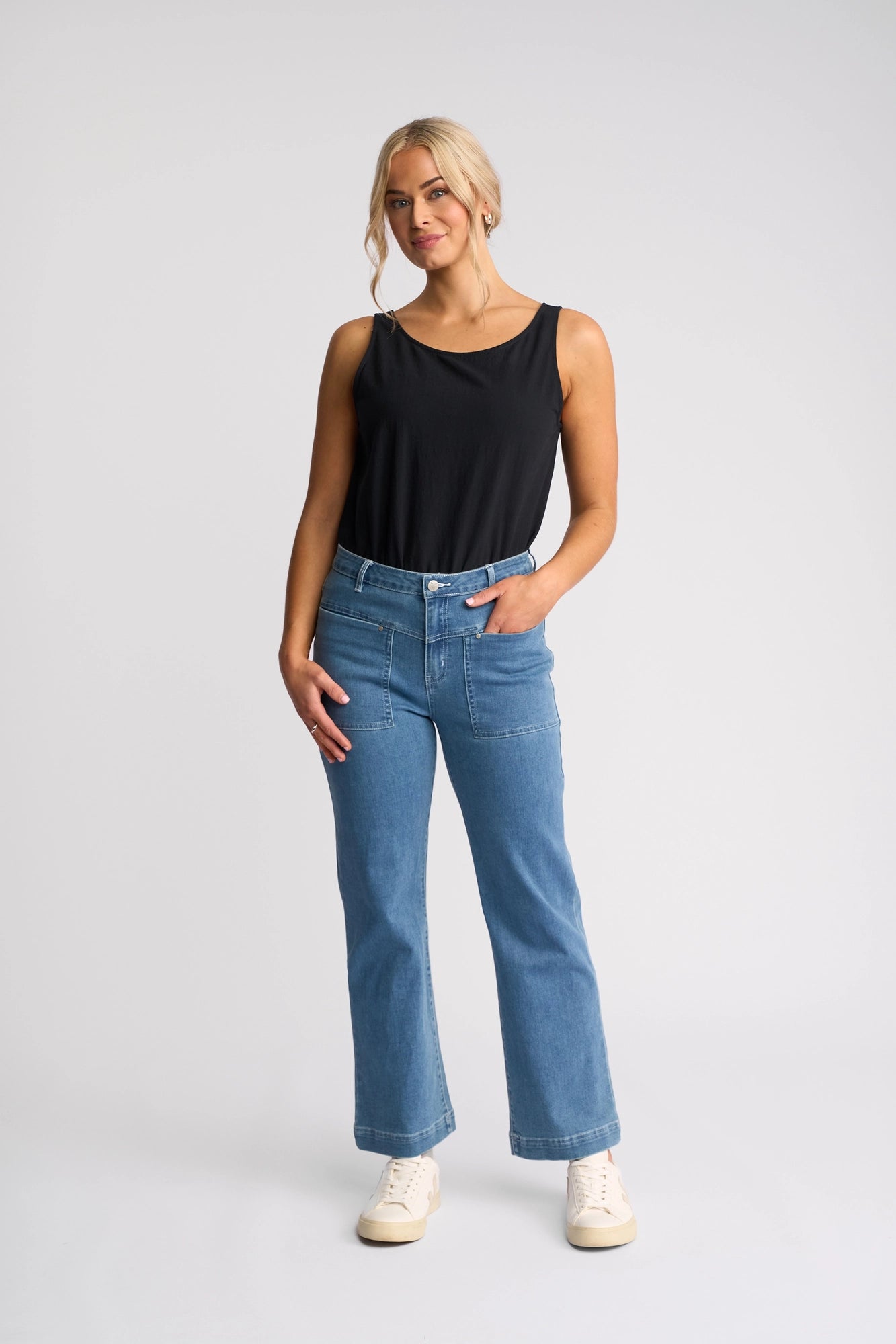A woman stands against a plain light background, wearing a sleeveless black top, white sneakers, and Vassalli’s Villa Jean in Blue Hue Denim. One hand is in her pocket and she has a relaxed expression.