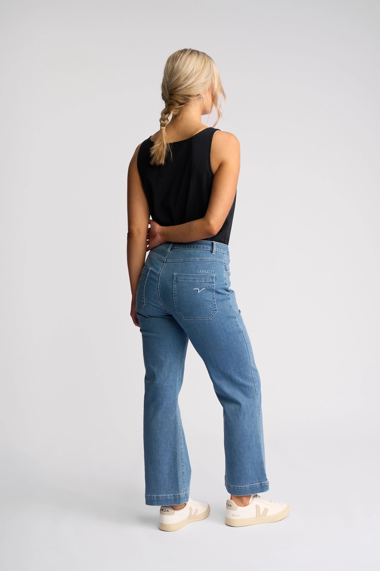 A woman with blonde hair in a braid wears a black sleeveless top, white sneakers, and the Villa Jean - Blue Hue Denim by Vassalli. She stands facing away, looking to her left against a plain white background.