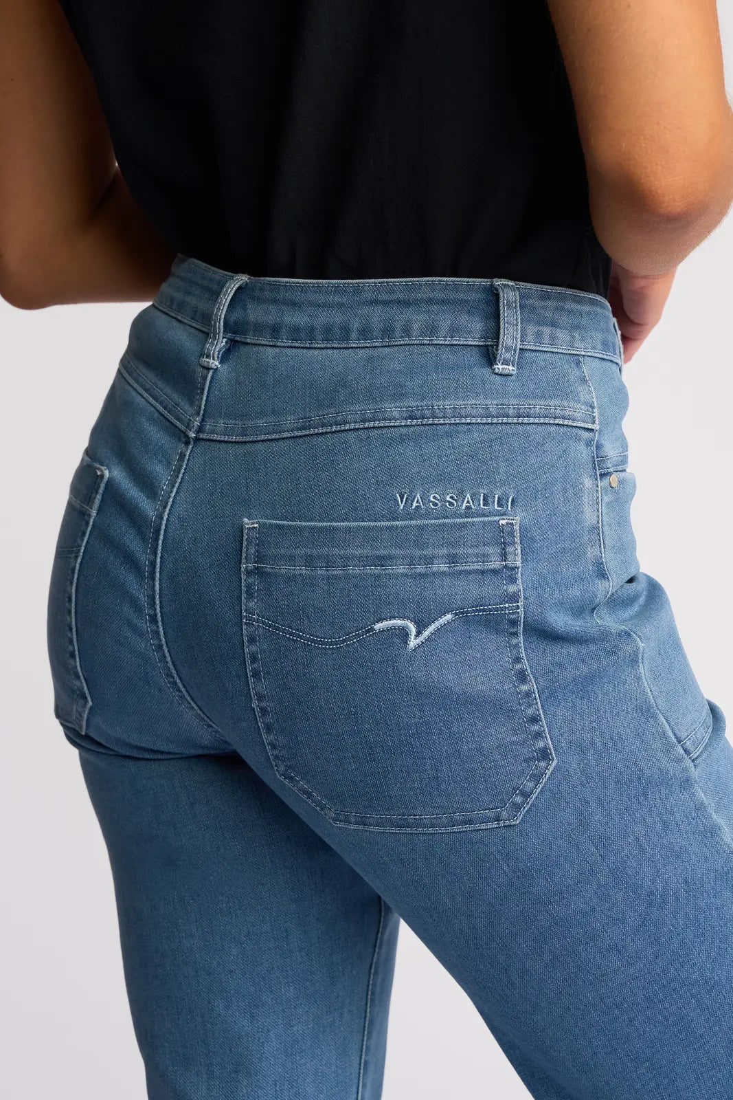 A person is shown wearing Vassalli’s Villa Jean in Blue Hue Denim, identifiable by the VASSALLI label above the back pocket and paired with a black top. Crafted from stretch denim for enhanced comfort and fit, only the lower back and upper legs are visible against a plain background.