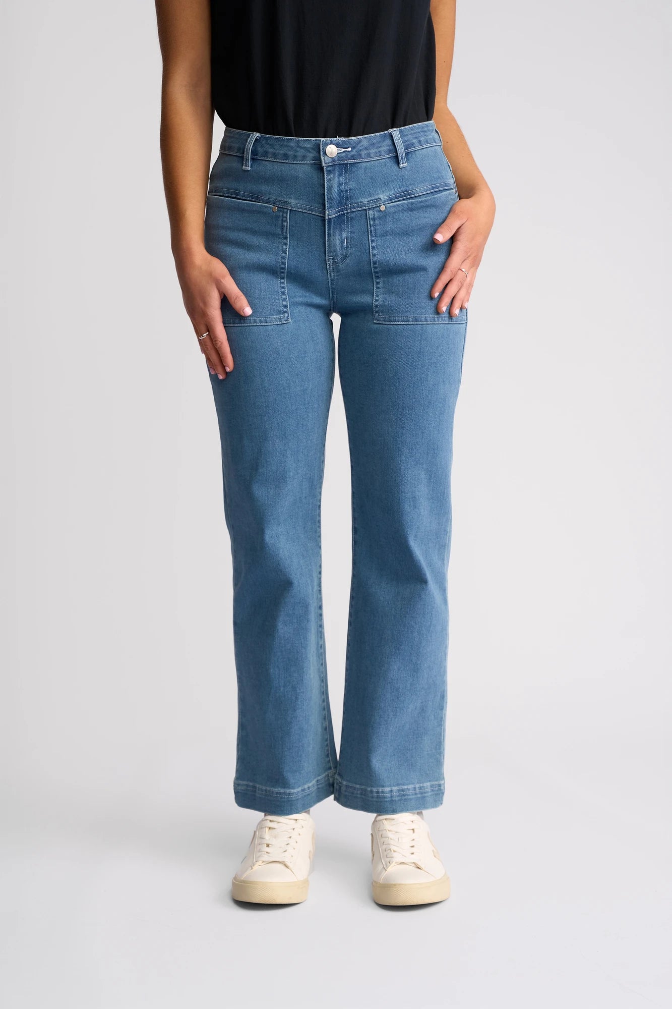 A person is shown from the shoulders down, standing against a plain light background, wearing Vassalli’s Villa Jean - Blue Hue Denim ankle-grazer jeans paired with a black top and white sneakers.