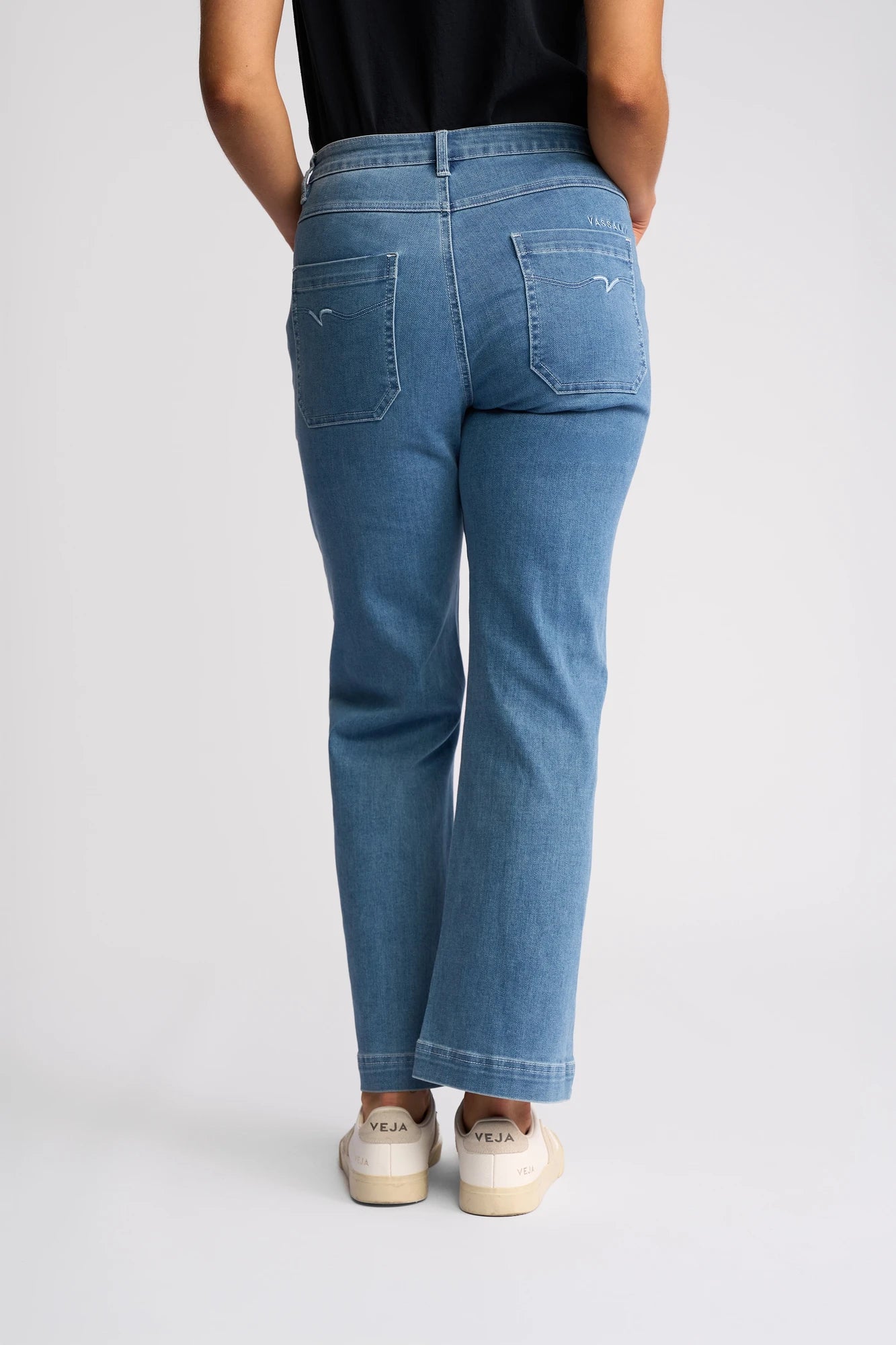 A person wearing the Vassalli Villa Jean in Blue Hue Denim, paired with a black top and white Veja sneakers, stands with their back to the camera against a plain light background.