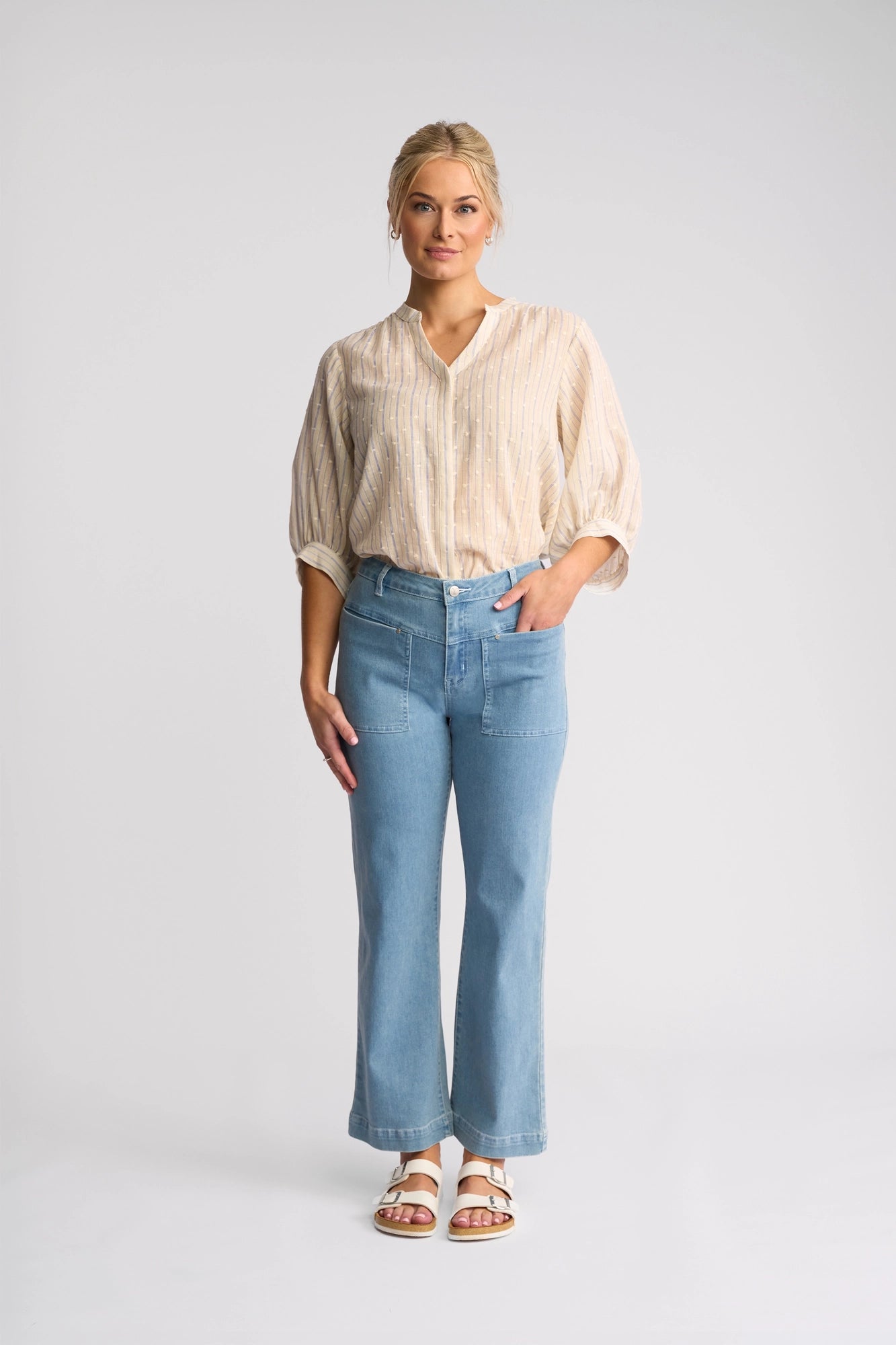 A woman stands against a plain background, wearing a beige striped blouse, white sandals, and the Vassalli Villa Jean in light wash denim. She has blonde hair pulled back and one hand in her pocket.