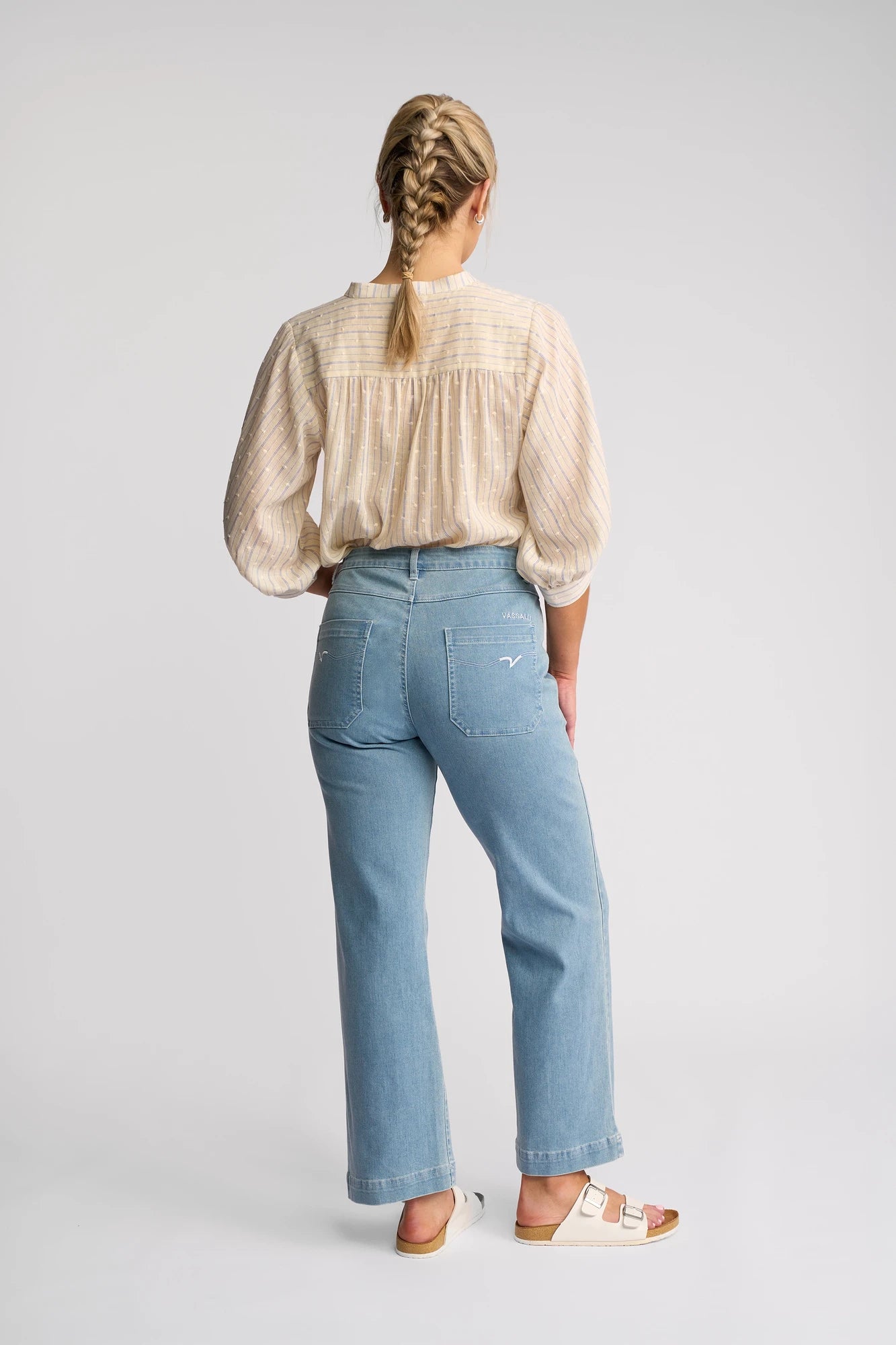 A person with braided hair, dressed in a beige long-sleeve blouse, Vassalli’s Villa Jean – Light Wash Denim, and white sandals stands facing away against a plain light background—the perfect look for summer jeans.