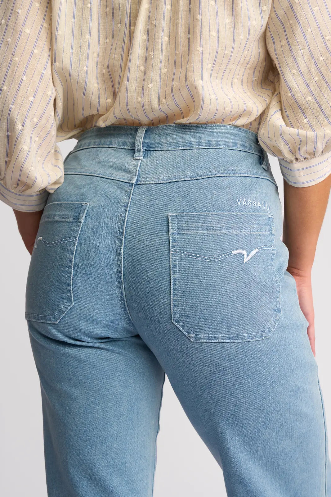 A person shown from the back is wearing the Vassalli Villa Jean in light wash denim, styled with straight legs and two rear pockets featuring white stitching and a small Vassalli logo. They have paired it with a beige, long-sleeve blouse adorned with blue stripes.
