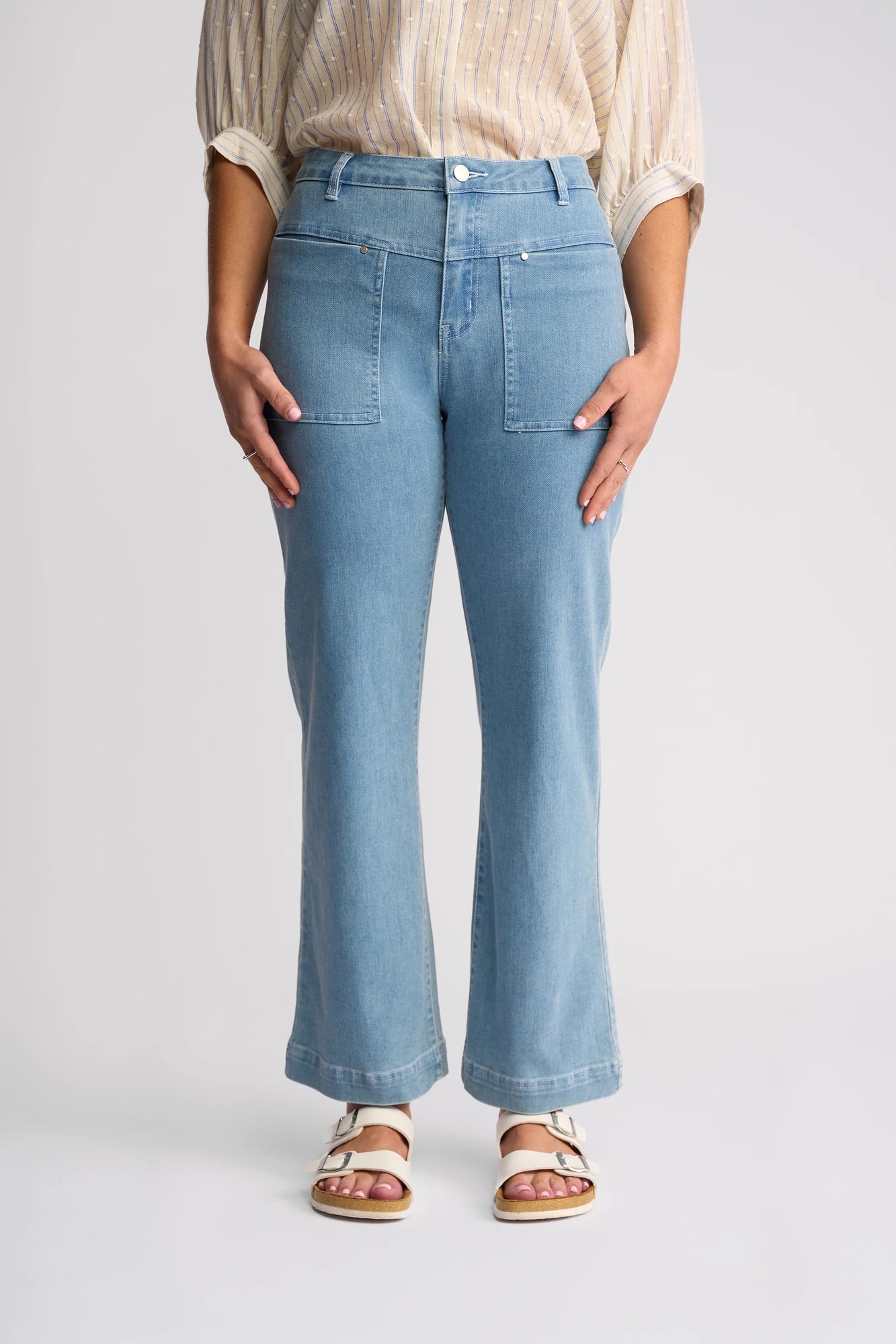 A woman is wearing Vassalli’s Villa Jean in Light Wash Denim, paired with a beige striped blouse and white sandals, standing against a plain light background.