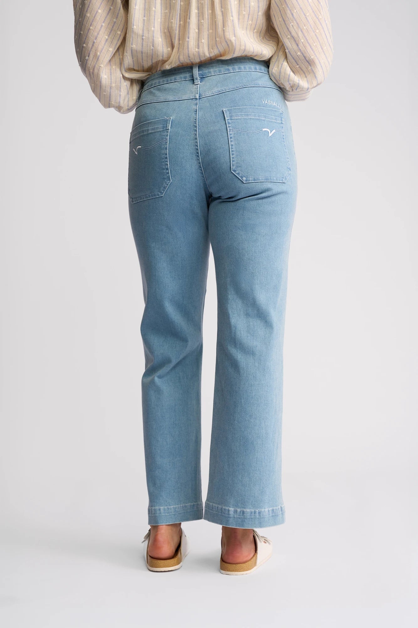 A person is photographed from the back against a plain white background, wearing the Vassalli Villa Jean - Light Wash Denim, which features a straight-leg fit, two back pockets, and is styled with flat shoes and a beige long-sleeve shirt.