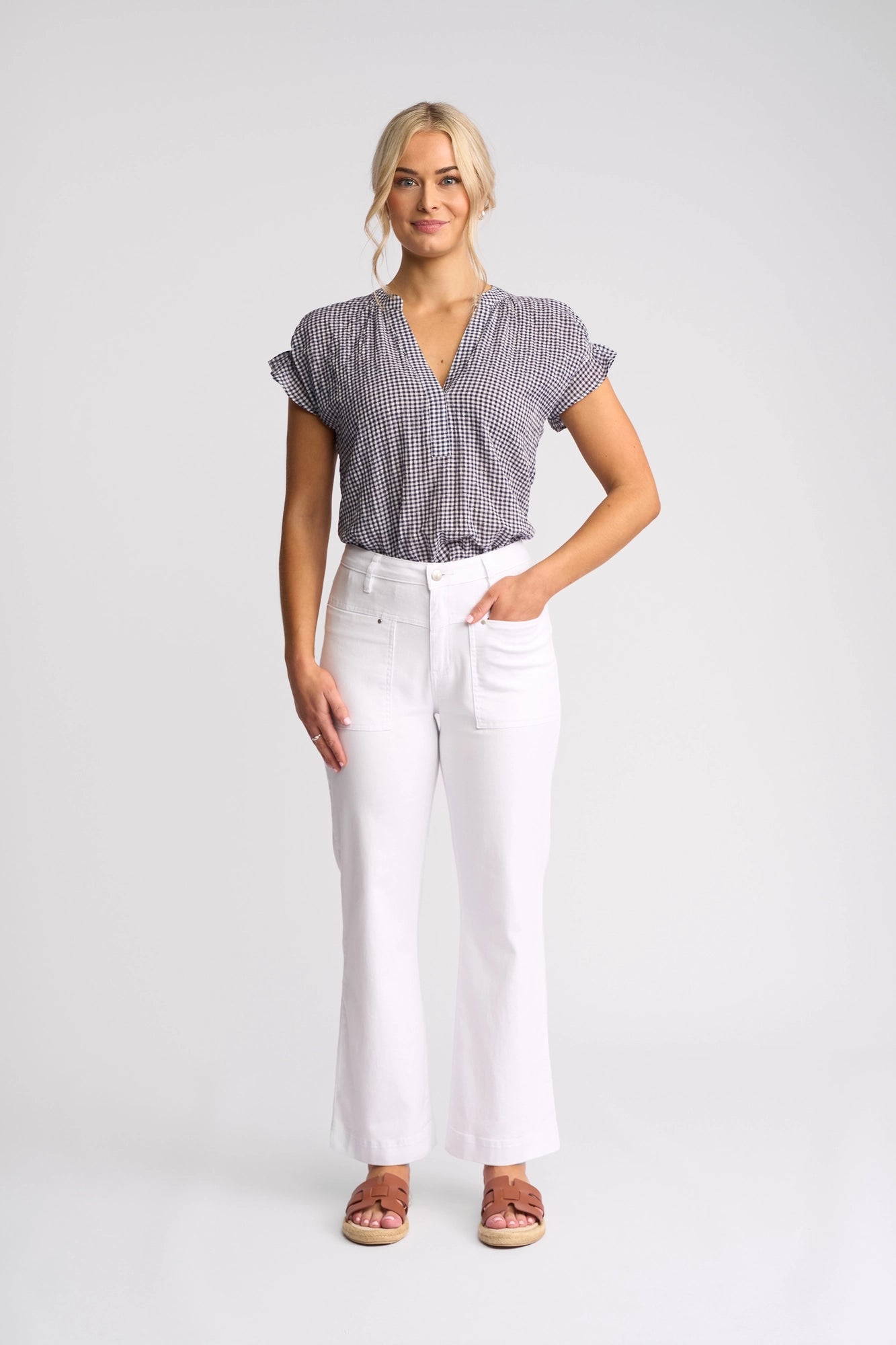 A woman stands against a plain background, smiling with one hand in her pocket, wearing the Vassalli Villa Jean in white denim—a high-waisted, straight-leg pant—paired with a black-and-white checkered blouse and brown sandals.