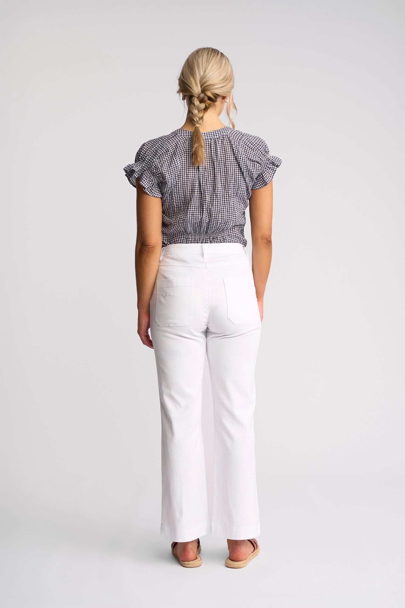 A woman with blonde hair in a braid stands facing away, wearing a short-sleeved checkered blouse, Vassalli Villa Jean - White Denim high-waisted straight-leg jeans, and sandals against a plain white background.