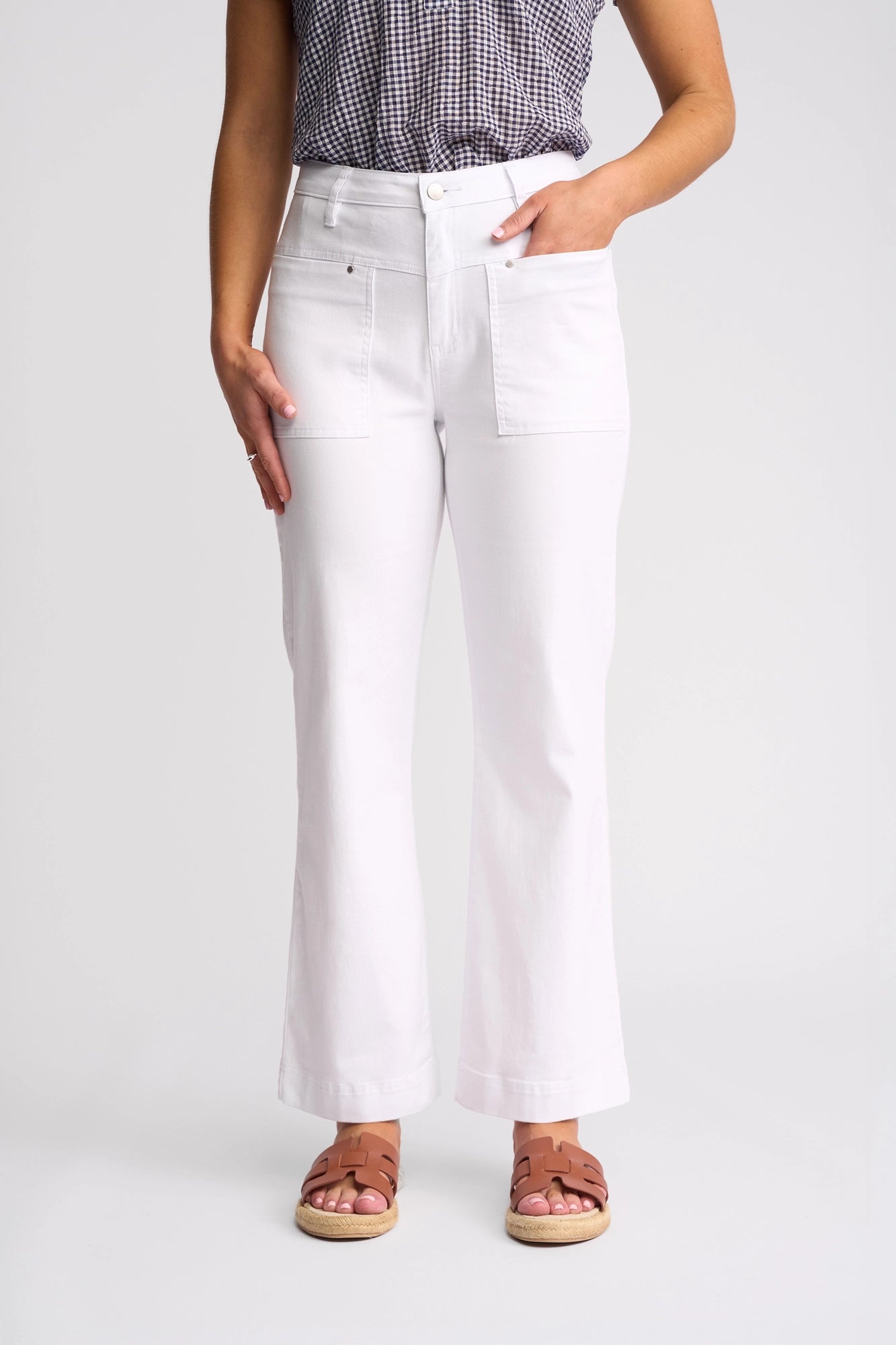 A person wearing Vassalli’s Villa Jean in white denim—a high-waisted, wide-leg summer style with front pockets—paired with a checked top and brown sandals stands against a plain background.
