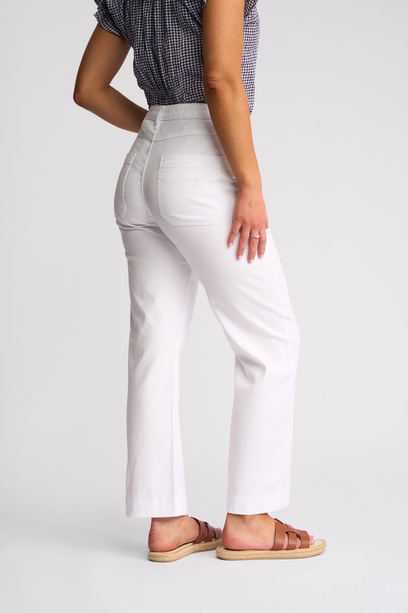 A person wearing a short-sleeve checkered top, brown sandals, and the Villa Jean - White Denim by Vassalli stands on a white background, facing away with one hand resting on their hip.