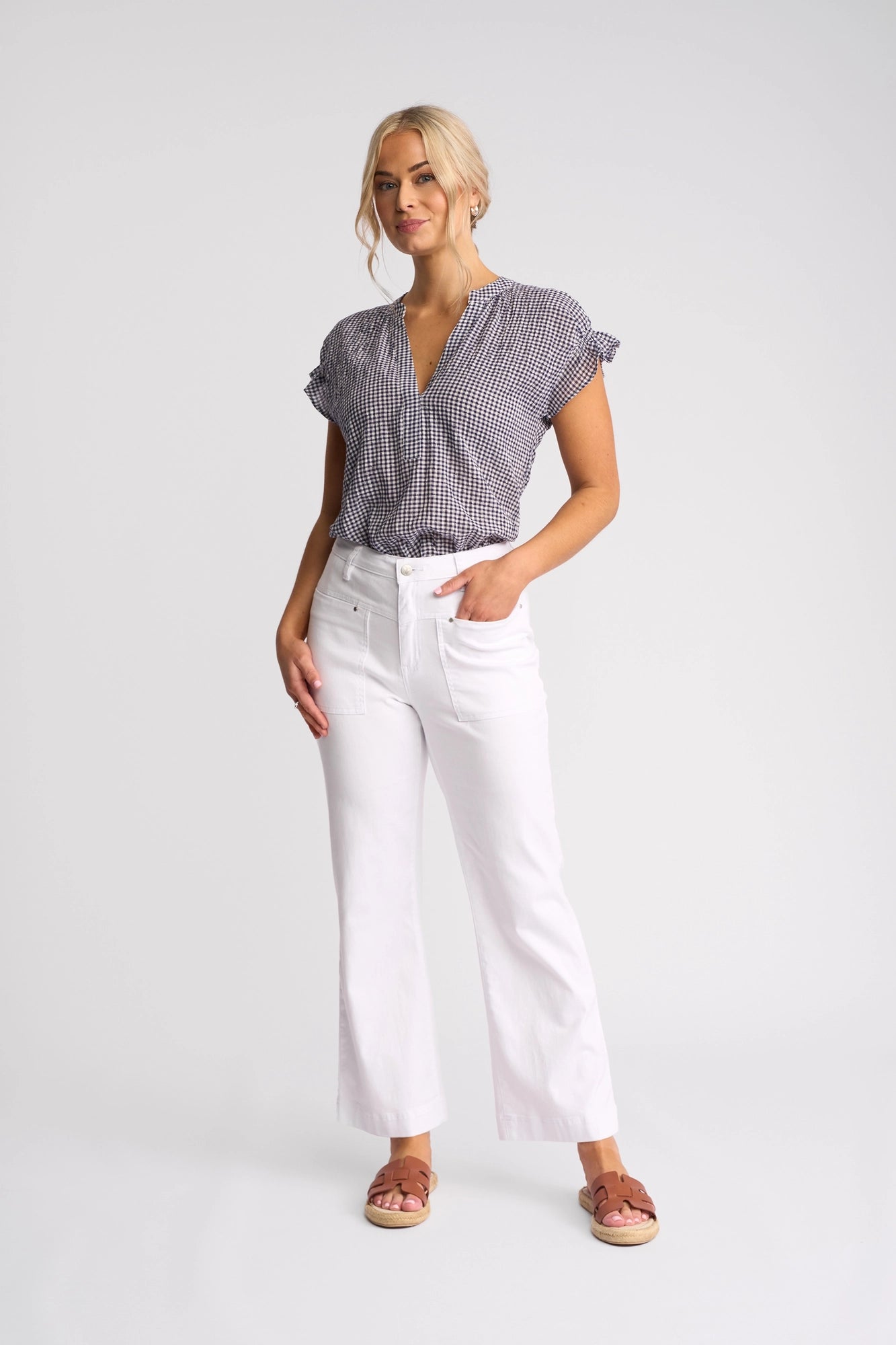 A woman with blonde hair styled in a loose updo poses against a plain background, wearing Vassalli’s Villa Jean – White Denim, paired with a short-sleeve checked blouse and sandals. She stands with one hand in her pocket.