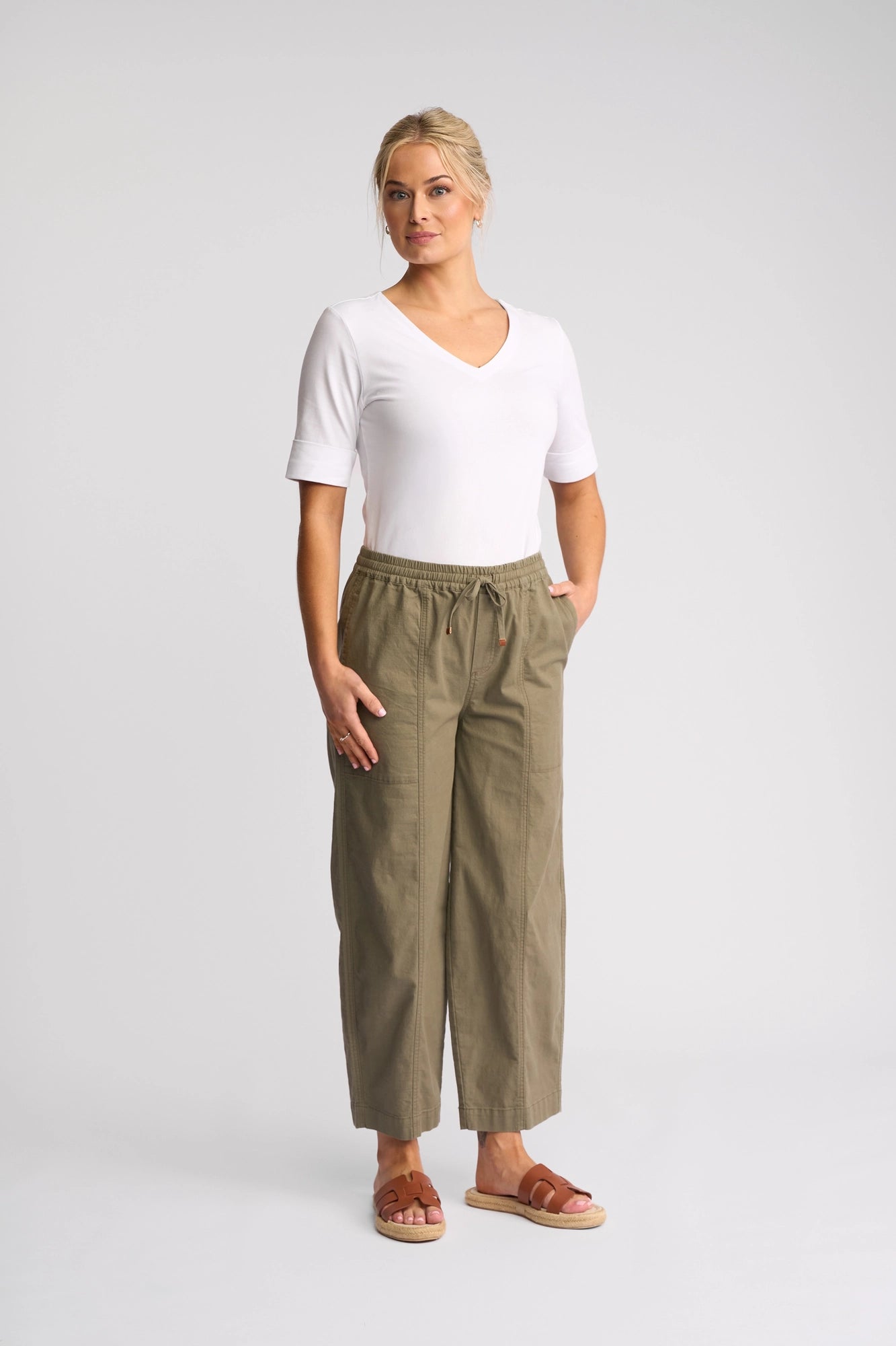 A blonde woman stands against a light grey background wearing a white v-neck t-shirt with rolled sleeves, the Vassalli Dione Pant in Army—a pair of loose-fitting, olive green ankle-grazer barrel-leg pants crafted from stretch cotton with a drawstring waist—and brown leather slide sandals.
