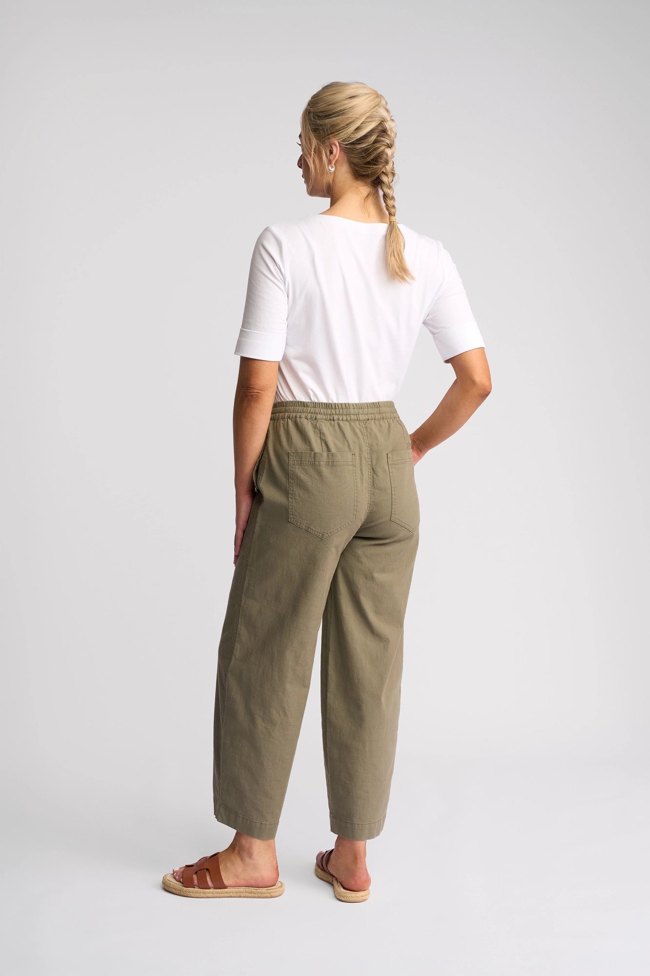 A woman with blonde hair in a braid is standing facing away, wearing a white t-shirt, Vassalli Dione Pant - Army, and brown sandals, posing against a plain light gray background.