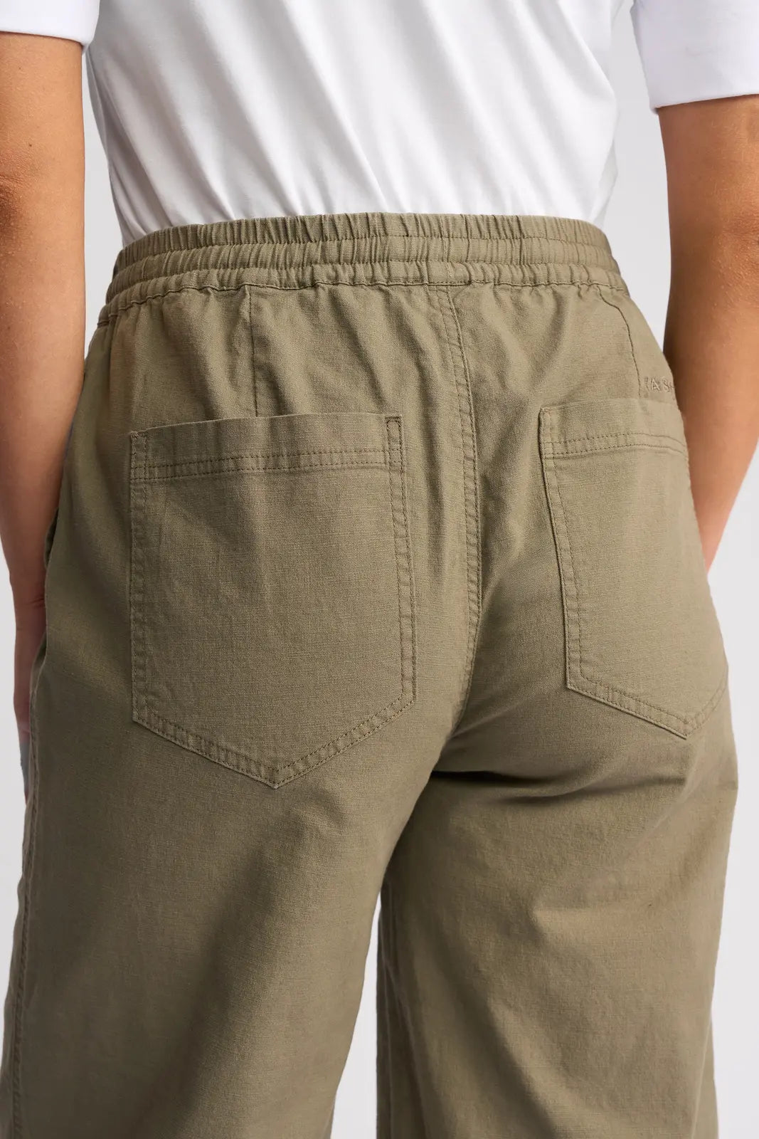 A person is wearing the Vassalli Dione Pant in Army, a pair of olive green, loose-fitting barrel-leg pants with back pockets, styled with a white shirt. The image displays the stretch cotton pants from the waist to mid-thigh, shown from behind.