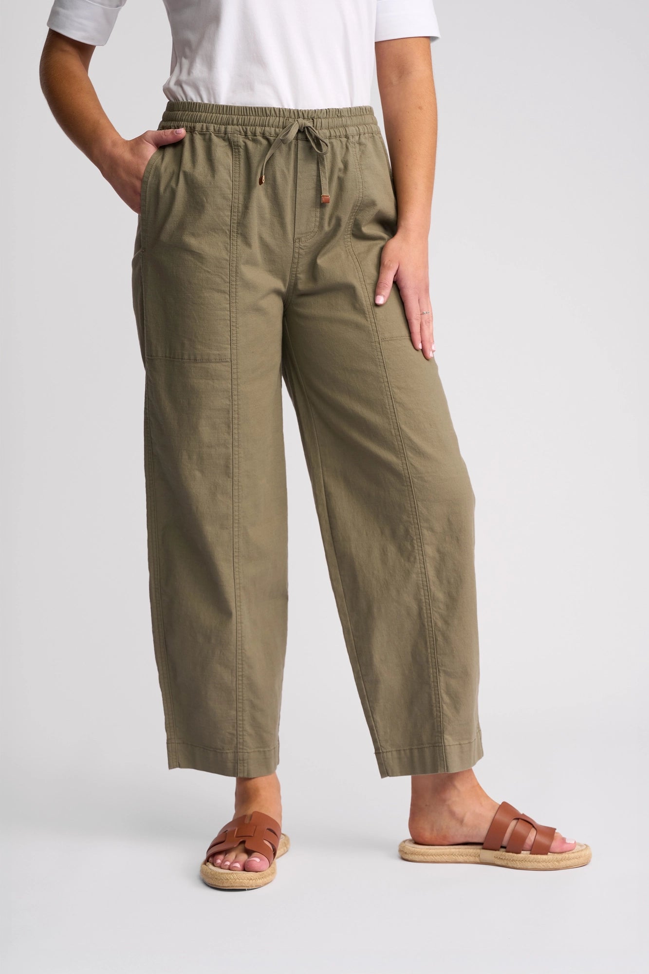 A person stands against a plain background with one hand in their pocket, wearing the Vassalli Dione Pant - Army (olive green stretch cotton drawstring pants), a white shirt, and brown sandals.