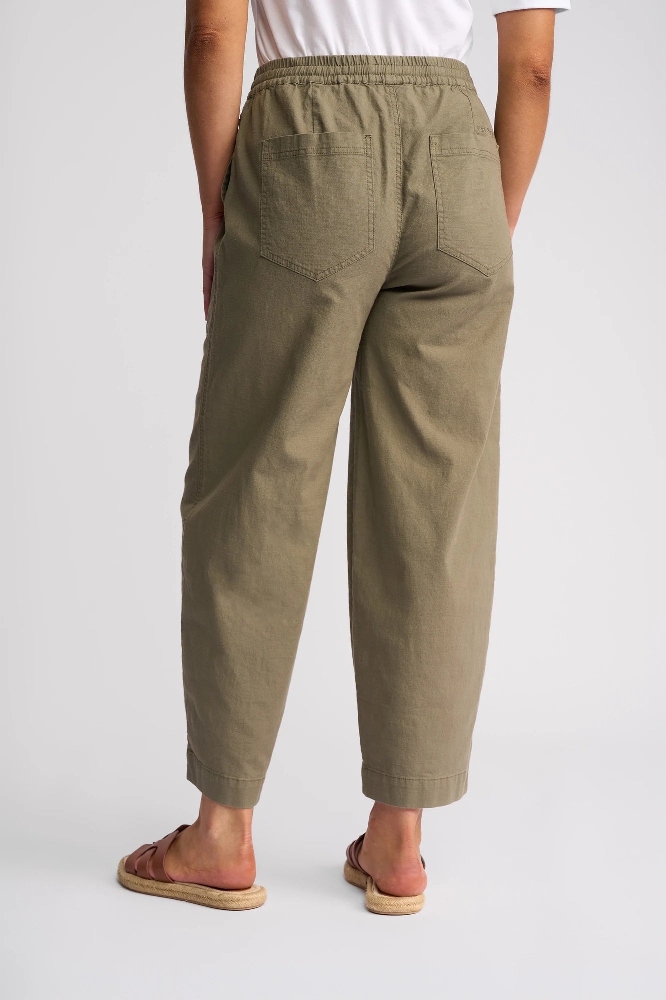 A person stands facing away on a plain light background, wearing Vassalli’s Dione Pant in Army—an olive green, high-waisted barrel-leg style with back pockets made from stretch cotton—paired with a white t-shirt and brown sandals.