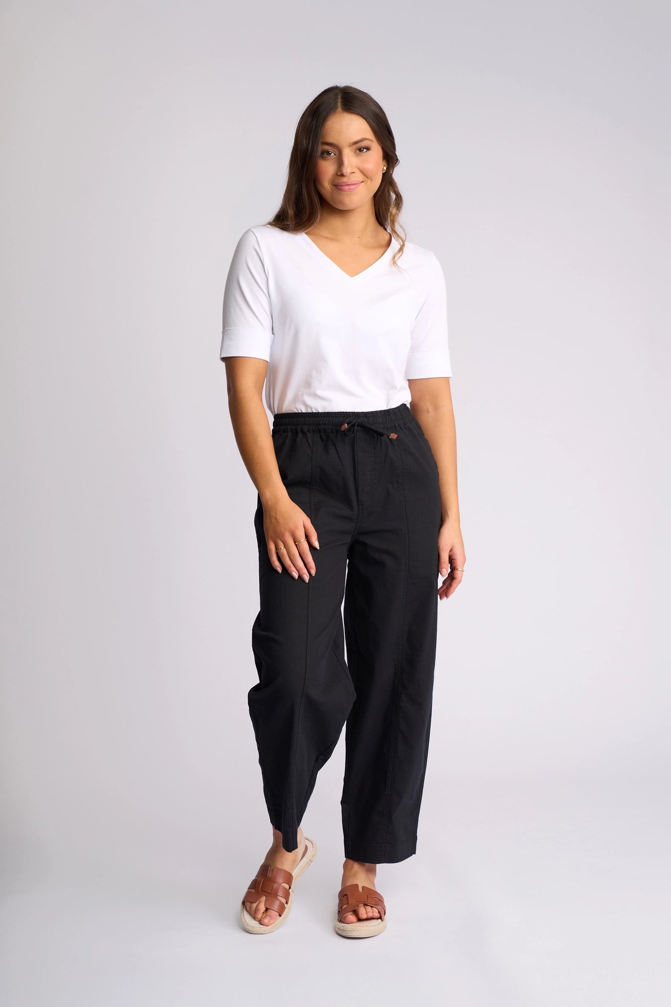 A woman stands against a plain background, wearing the Vassalli Dione Pant in black, featuring an elasticated drawstring waistband, paired with a white V-neck T-shirt and brown sandals. She has long brown hair and poses casually with a relaxed demeanor and a slight smile.
