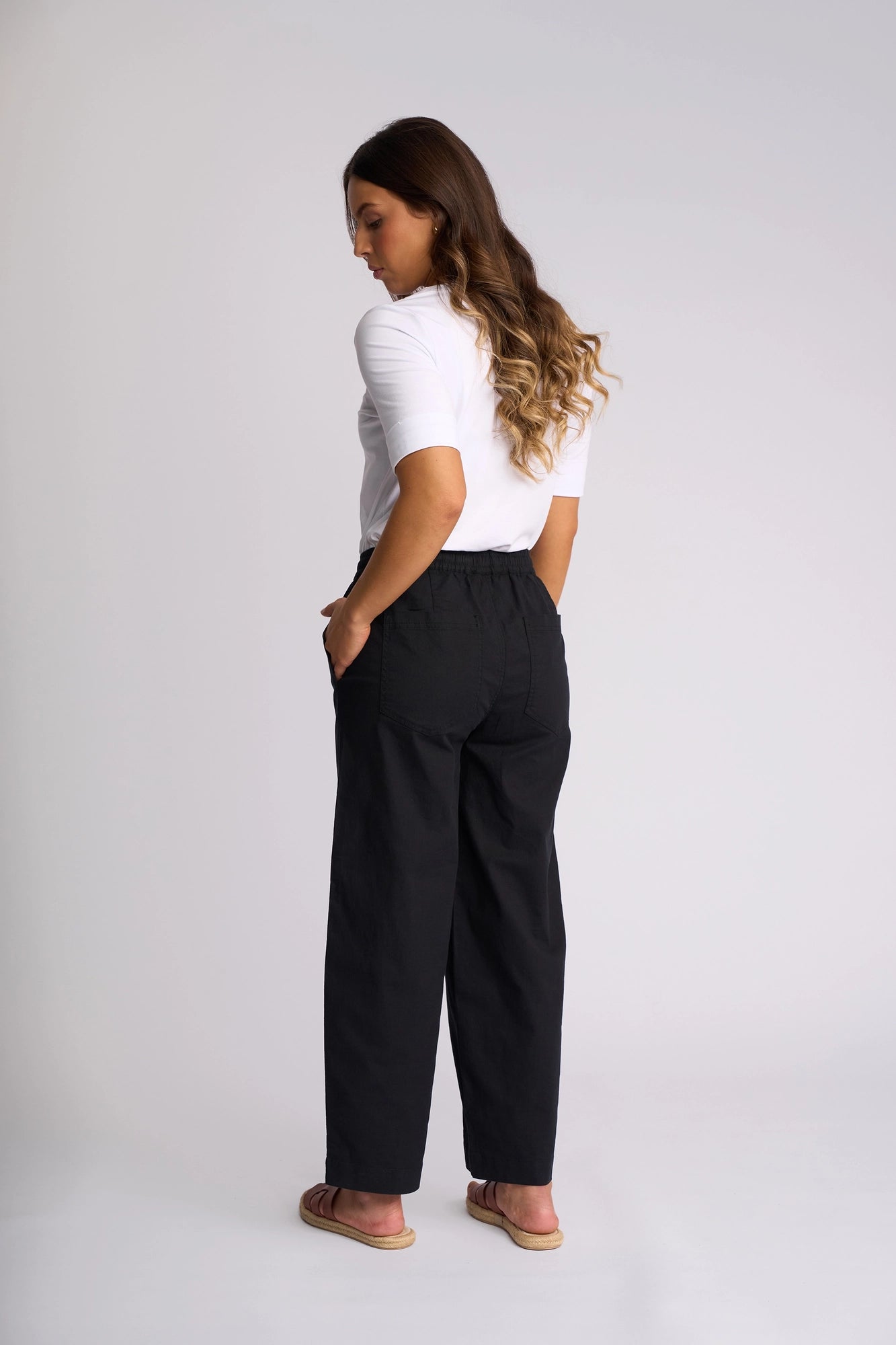 A woman with long wavy hair stands against a plain light background, facing slightly away with her hands in her pockets. She wears a white short-sleeve top paired with the Vassalli Dione Pant in black—high-waisted barrel-leg pants crafted from stretch cotton and featuring an elasticated drawstring waistband. Sandals complete this relaxed yet polished look.