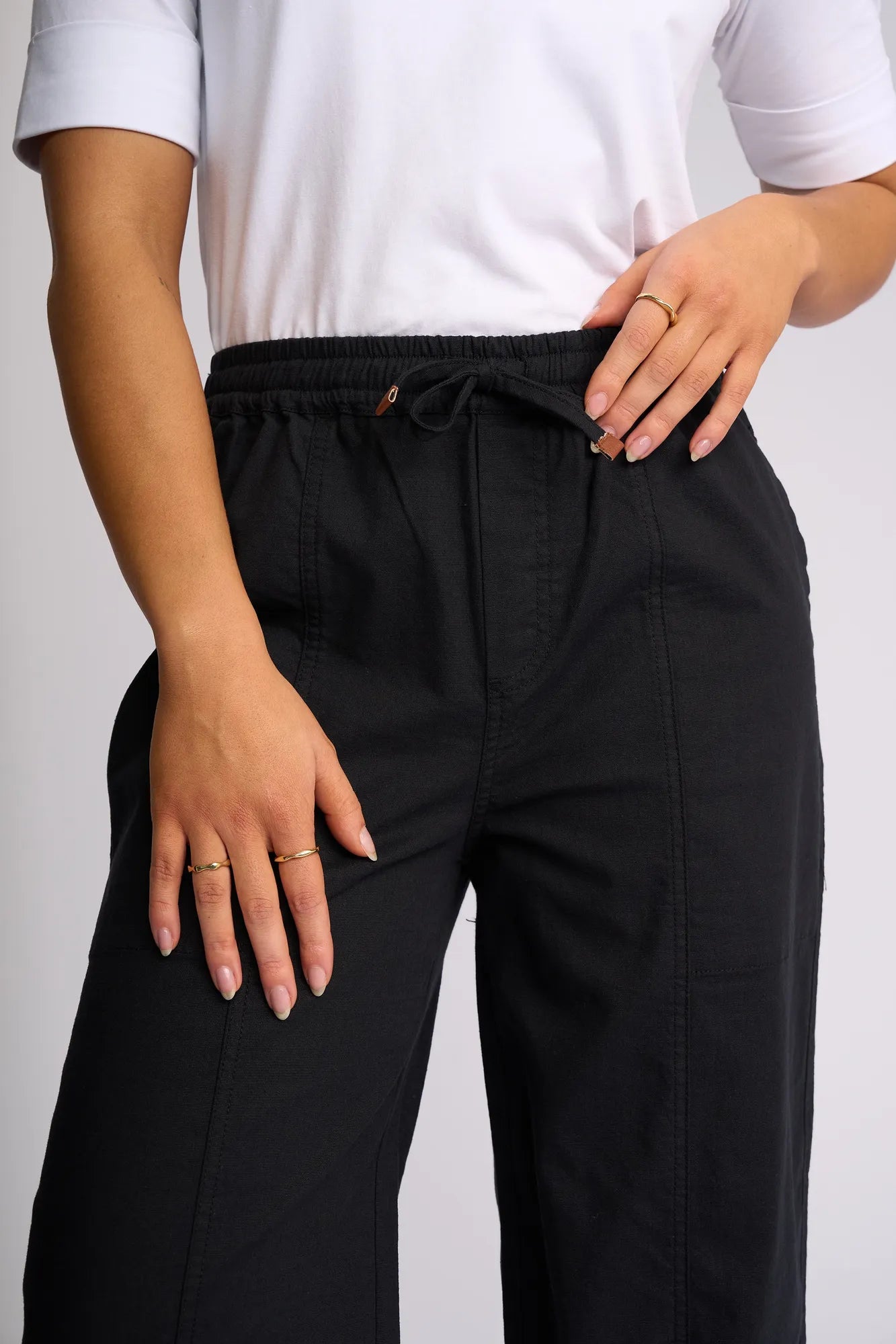 A person is wearing a white shirt paired with the Vassalli Dione Pant in black, which features a barrel-leg silhouette and an elasticated drawstring waistband. They stand with one hand resting on their hip and the other by their side, displaying gold rings on their fingers.
