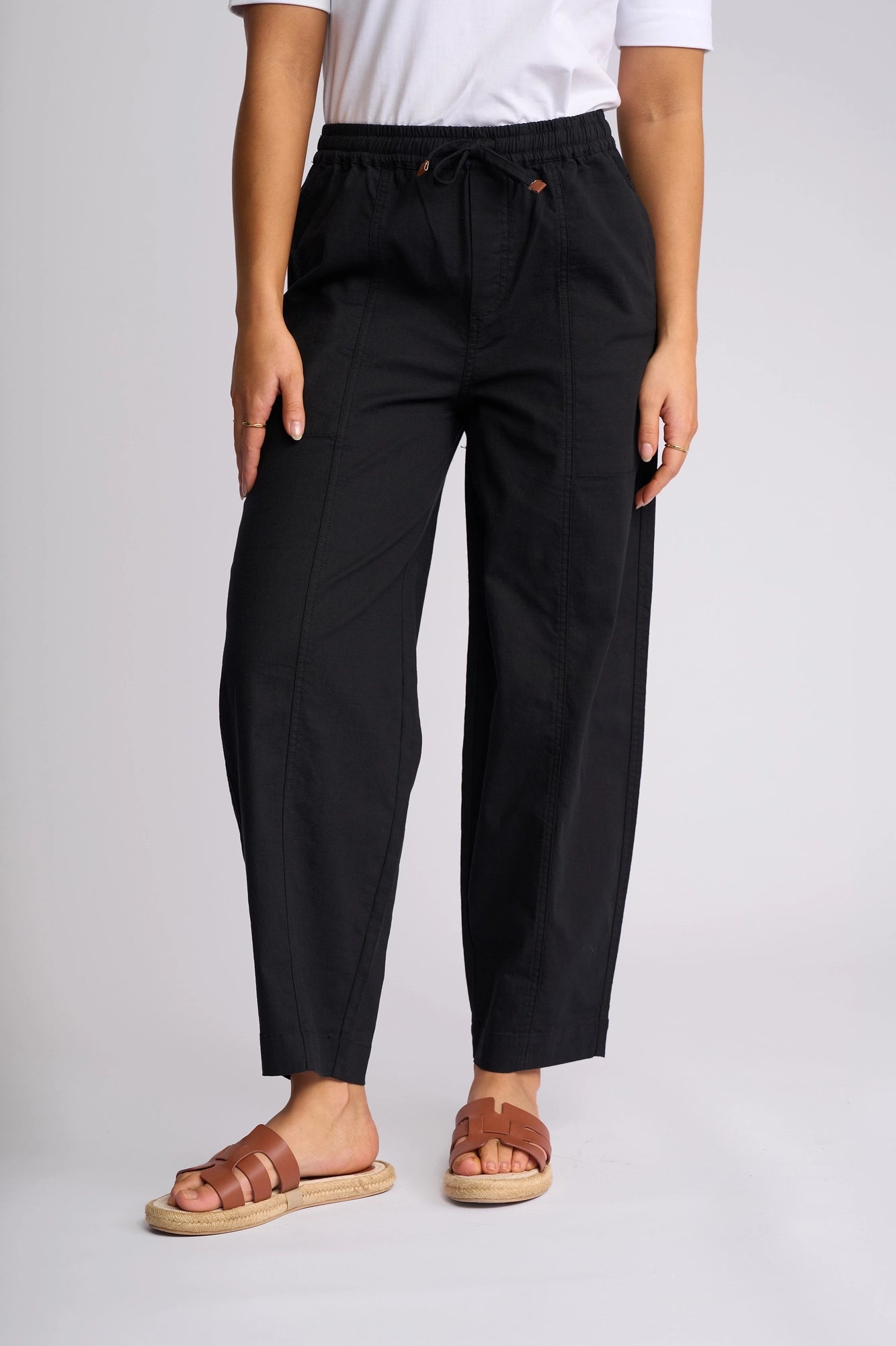 A person is shown from the waist down wearing Vassalli’s Dione Pant in black, featuring an elasticated drawstring waistband, paired with a white shirt and brown sandals against a plain light background.
