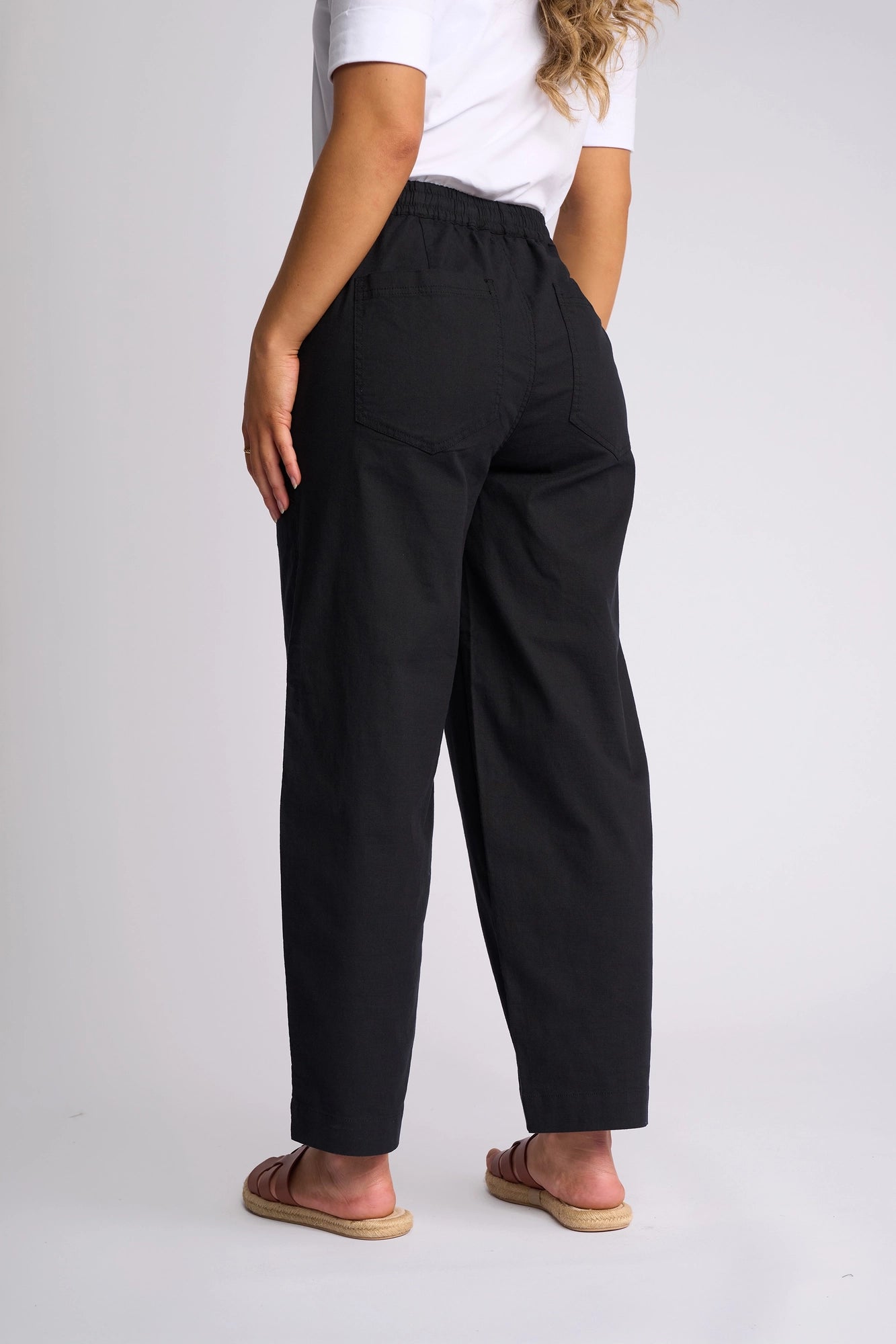A person stands facing away, wearing a white t-shirt and the Vassalli Dione Pant - Black, featuring back pockets and an elasticated drawstring waistband. Crafted from stretch cotton for added comfort and ease, these barrel-leg pants are paired with sandals to complete the look.