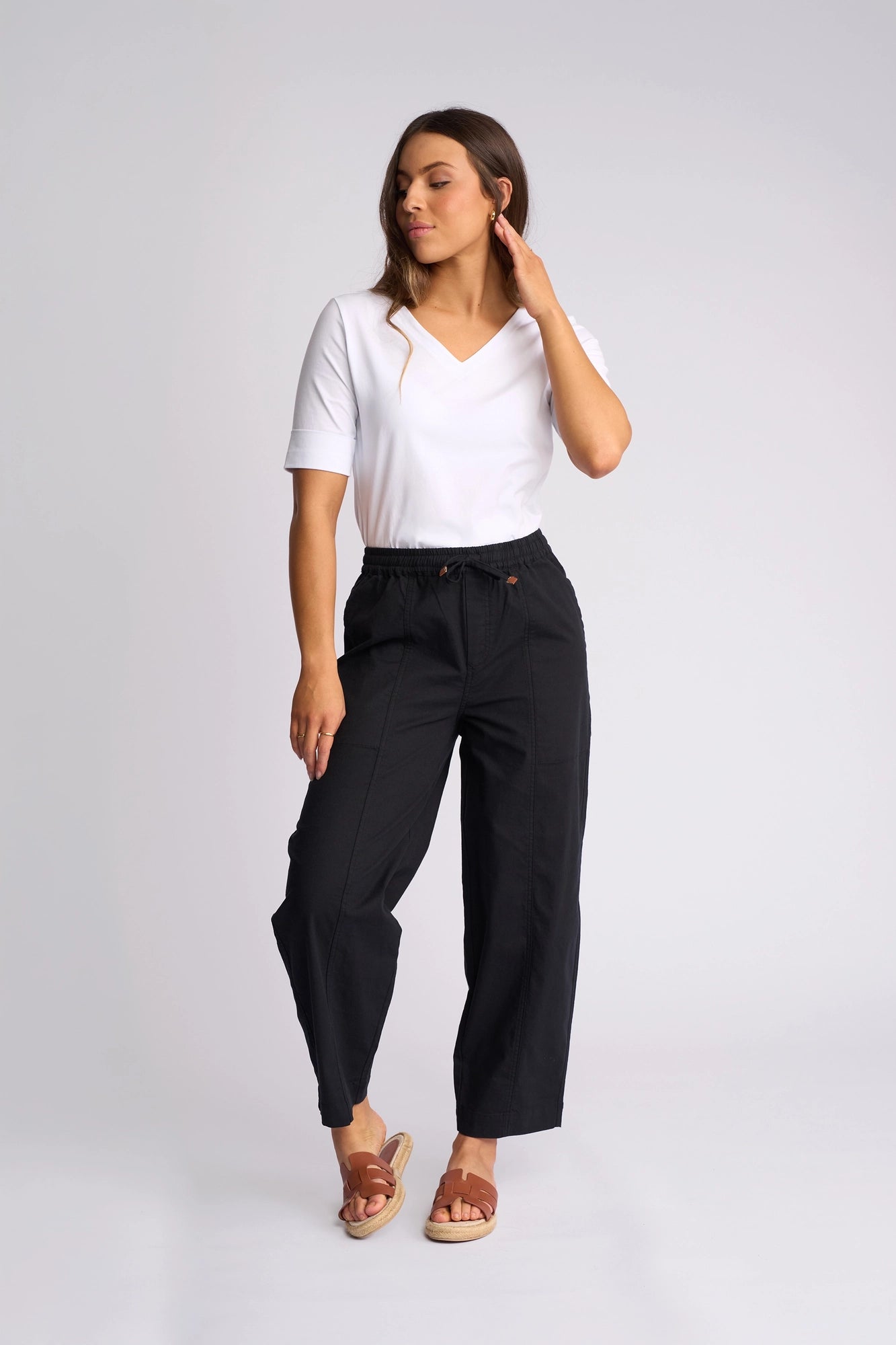 A woman stands against a plain light background, touching her hair and looking to the side, dressed in a white V-neck T-shirt, sandals, and the Vassalli Dione Pant in black with its high-waisted barrel-leg silhouette and elasticated drawstring waistband.
