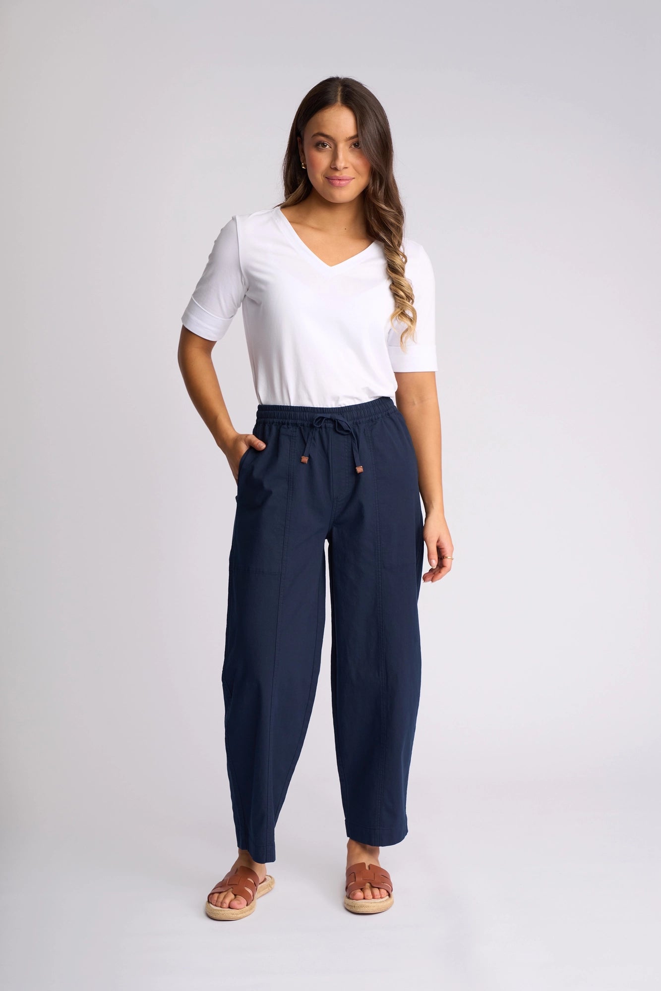 A woman stands against a plain background wearing a white short-sleeve V-neck shirt paired with the Vassalli Dione Pant in Ink. These high-waisted, barrel-leg pants are crafted from soft stretch cotton and feature an ankle-grazer length with a drawstring waist. She poses with one hand in her pocket, exuding a relaxed expression, and completes the look with brown sandals.