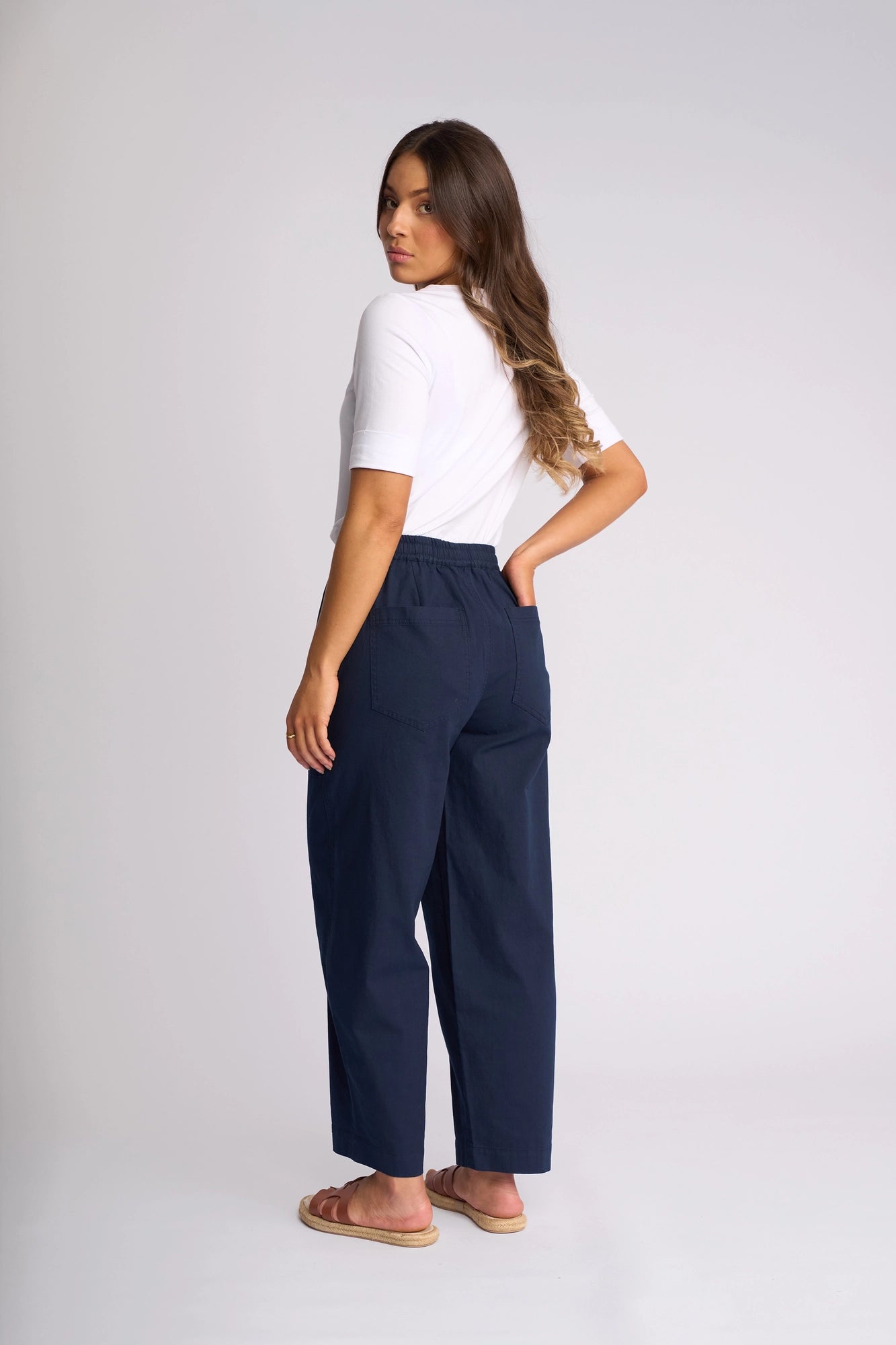 A woman stands sideways, looking over her shoulder. She wears a white short-sleeve top paired with the Vassalli Dione Pant in Ink—navy blue barrel-leg ankle-grazer pants with pockets, crafted from stretch cotton. Tan sandals complete her look. Her long hair is down, and the background is plain light grey.