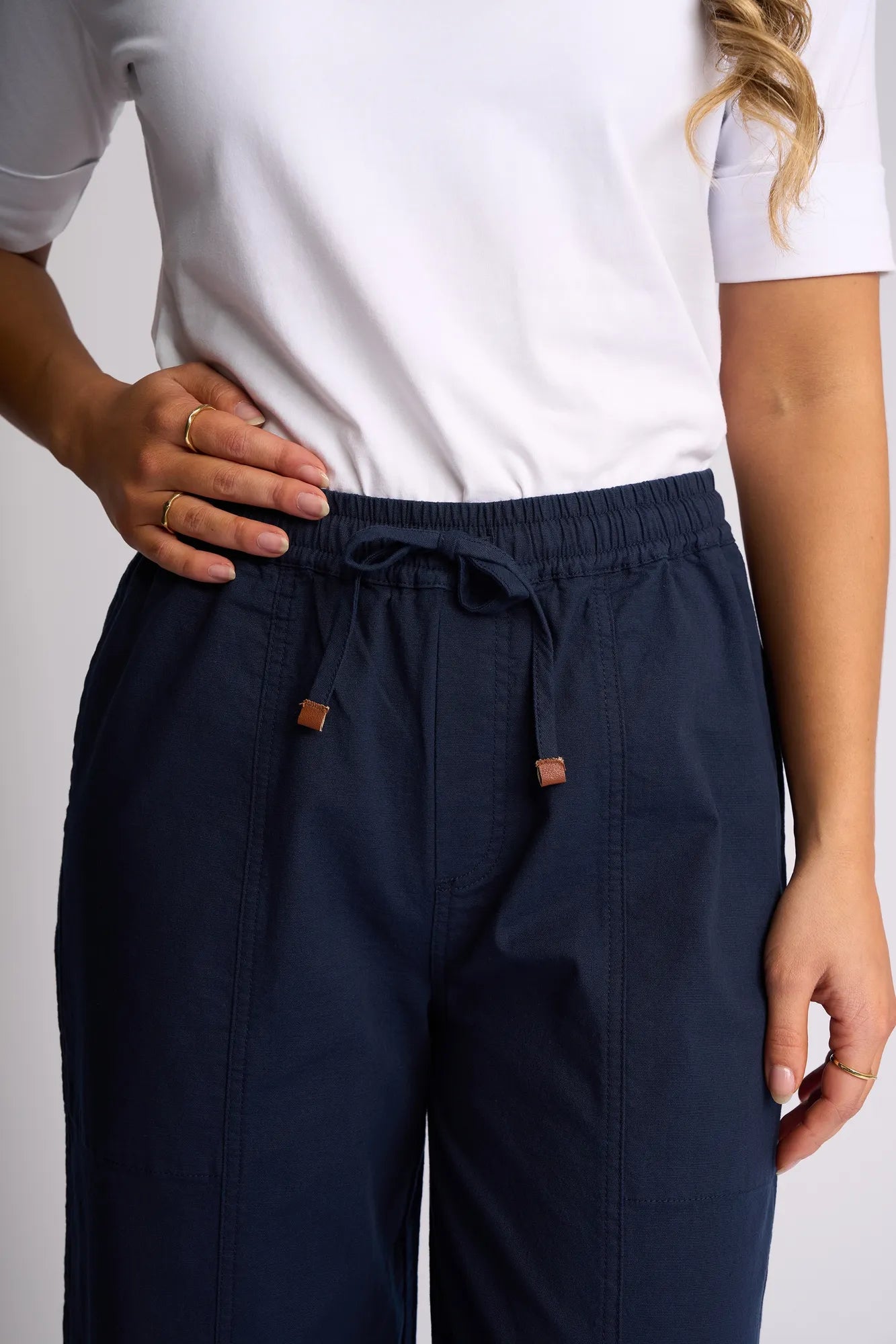 A person wearing a white short-sleeve shirt and the Vassalli Dione Pant in Ink—an ankle-grazer barrel-leg style in stretch cotton—stands with one hand on their waist and the other relaxed at their side.