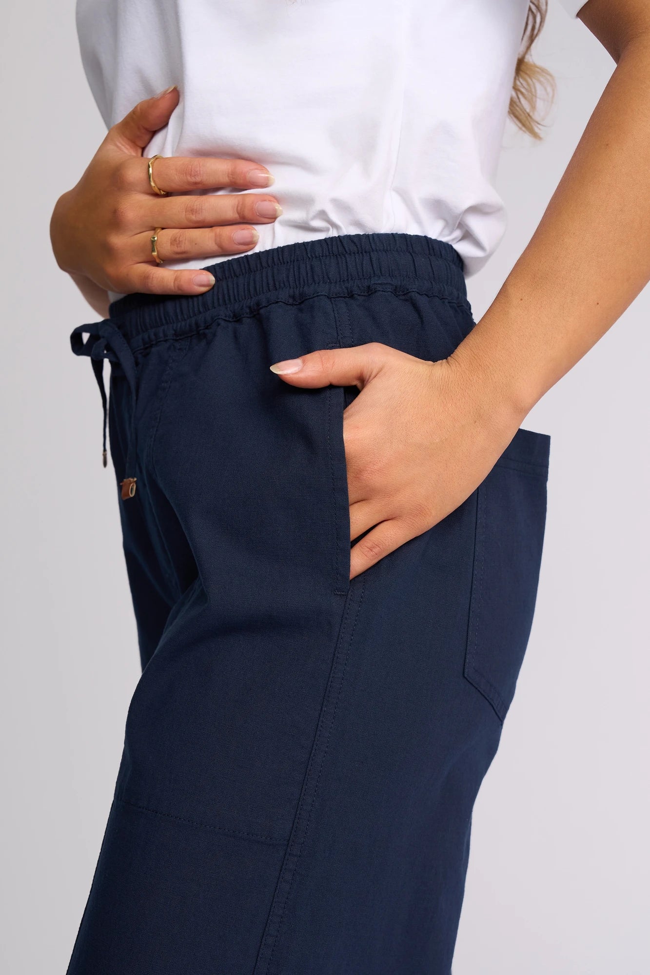 A person wearing a white shirt and Vassalli Dione Pant - Ink stands with one hand in their pocket and the other resting on their waist.