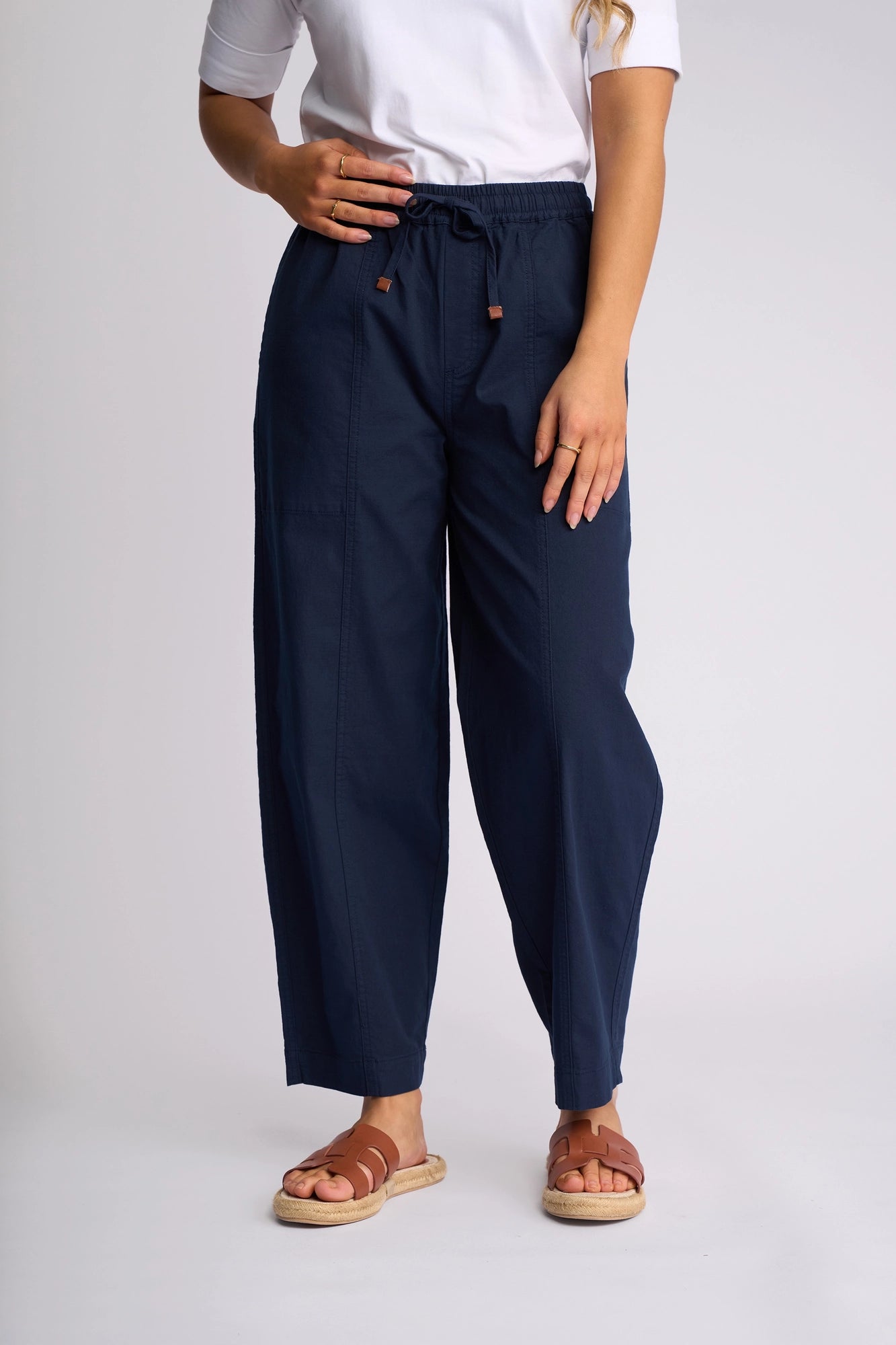 A person wearing a white top, the Vassalli Dione Pant in Ink—an ankle-grazer barrel-leg style crafted from stretch cotton—and brown sandals stands against a plain light background. Only the lower half of their body is visible.