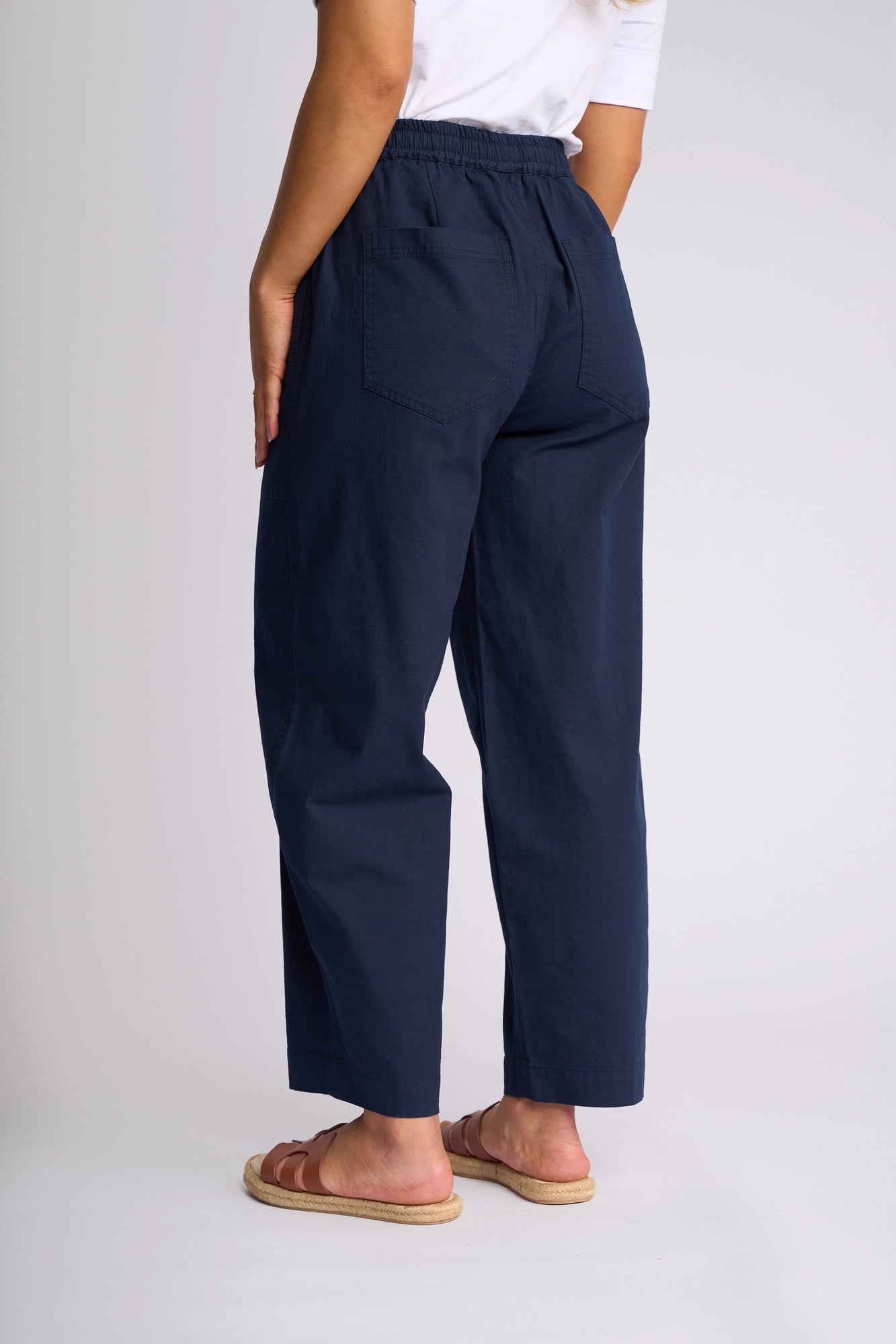 A person stands facing away, wearing a white shirt and the Vassalli Dione Pant in Ink—navy blue high-waisted barrel-leg pants crafted from stretch cotton with back pockets—paired with brown sandals against a plain background.