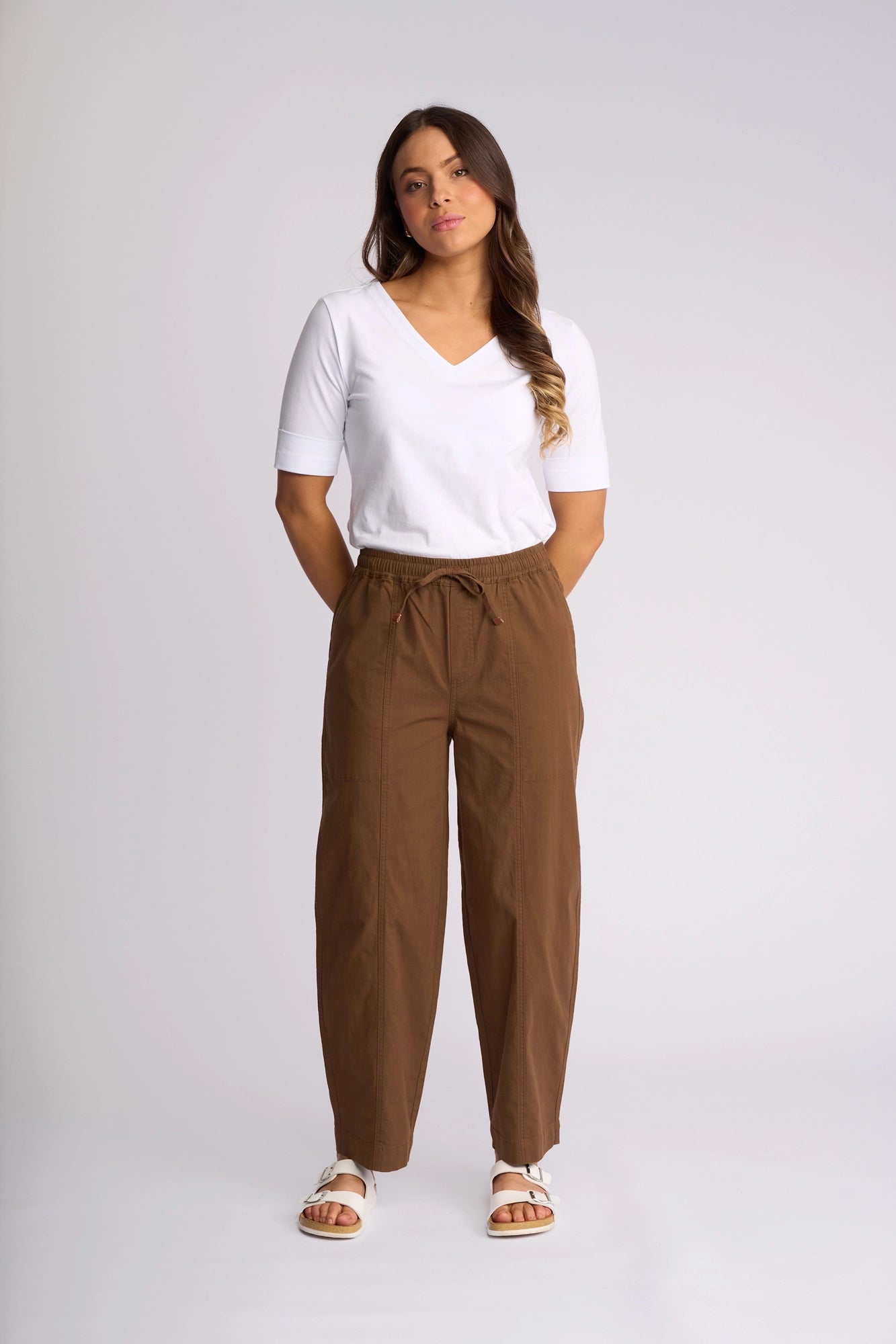 A woman stands against a plain background, wearing a white V-neck top, the Vassalli Dione Pant in Pecan—a loose barrel-leg style with an elasticated drawstring waistband—and white sandals. Her hands are in her pockets, and she has long brown hair.