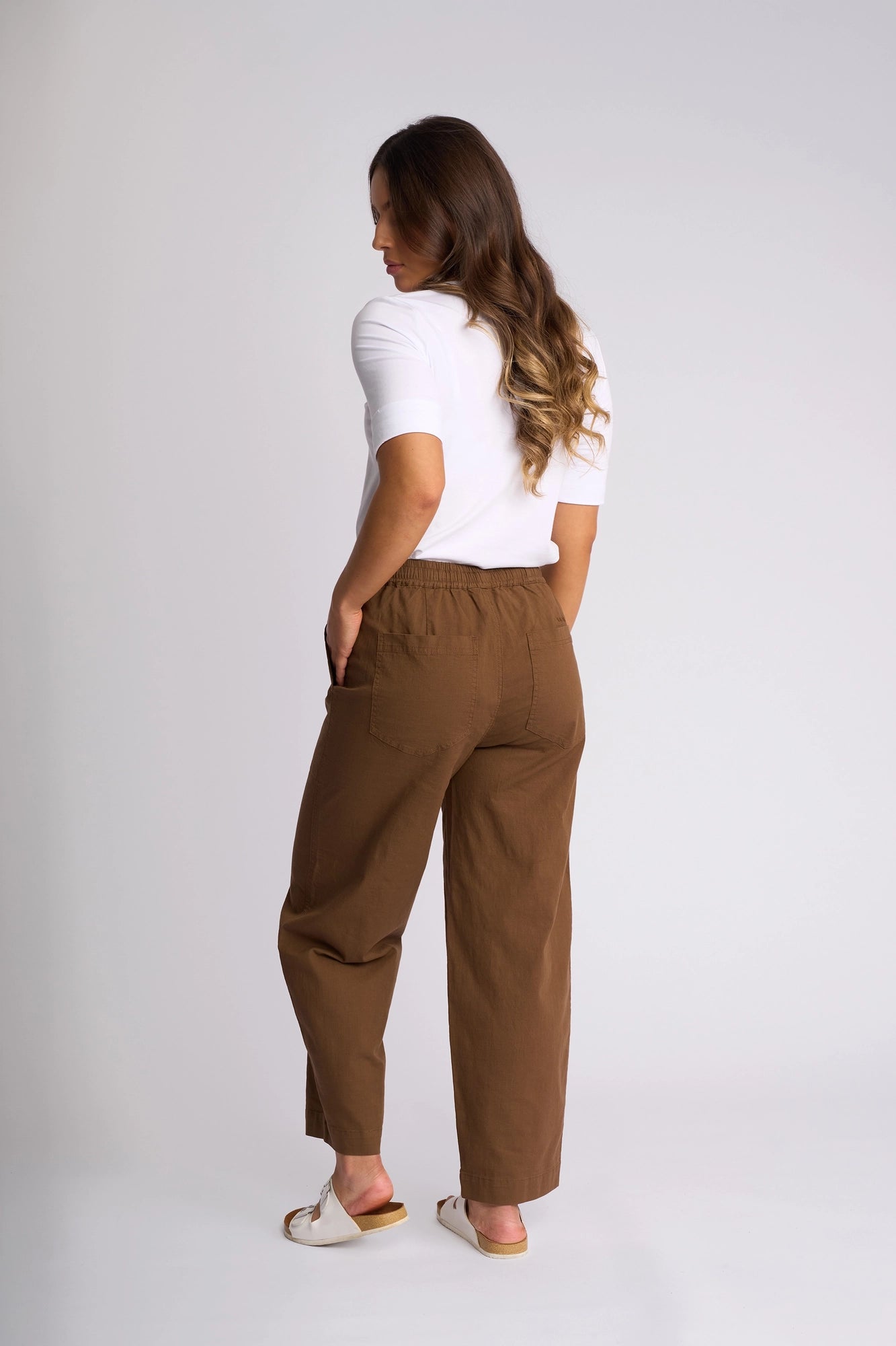 A woman stands against a plain background, facing away. She wears a white shirt styled with the Vassalli Dione Pant in Pecan—brown barrel-leg pants featuring an elasticated drawstring waistband and crafted from stretch cotton for comfort. White sandals complete her look as her hands rest in her pockets and her long wavy hair falls over her shoulder.