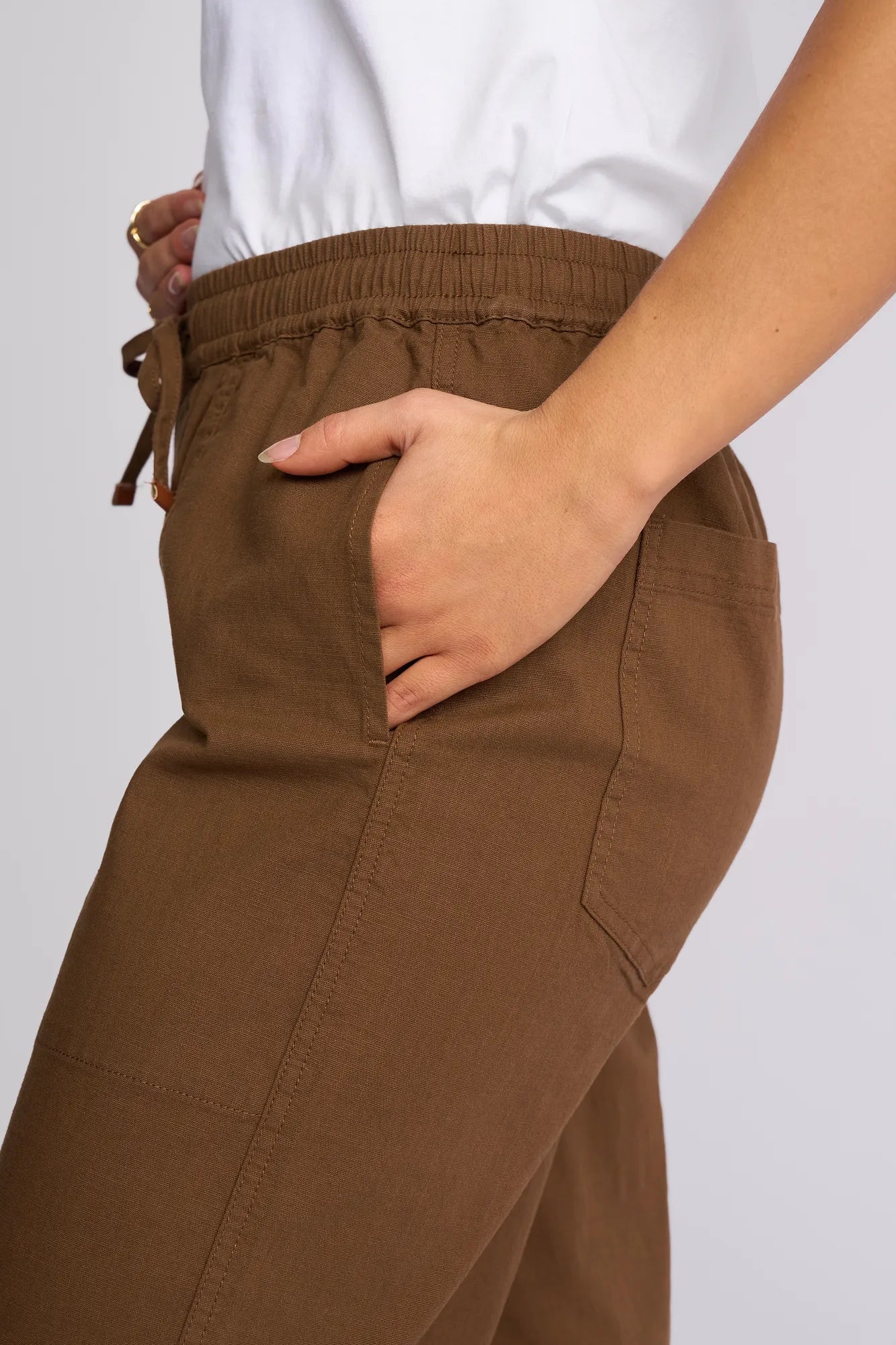 A person wearing the Vassalli Dione Pant in Pecan, featuring an elasticated drawstring waistband, and a white shirt has their right hand in their pocket. Only the side of their torso and upper legs are visible against a plain background.