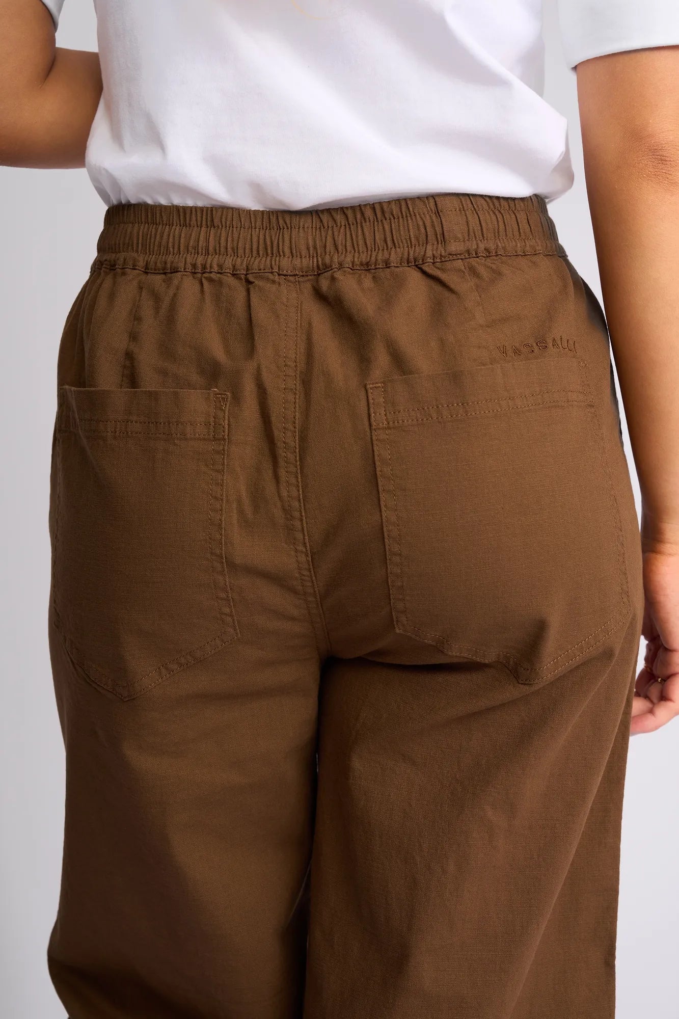 A person is shown from the back wearing the Vassalli Dione Pant in Pecan, paired with a white shirt. The barrel-leg pants are crafted from stretch cotton and feature an elasticated drawstring waistband and two back pockets. Only part of the wearer’s arm is visible on the right side.