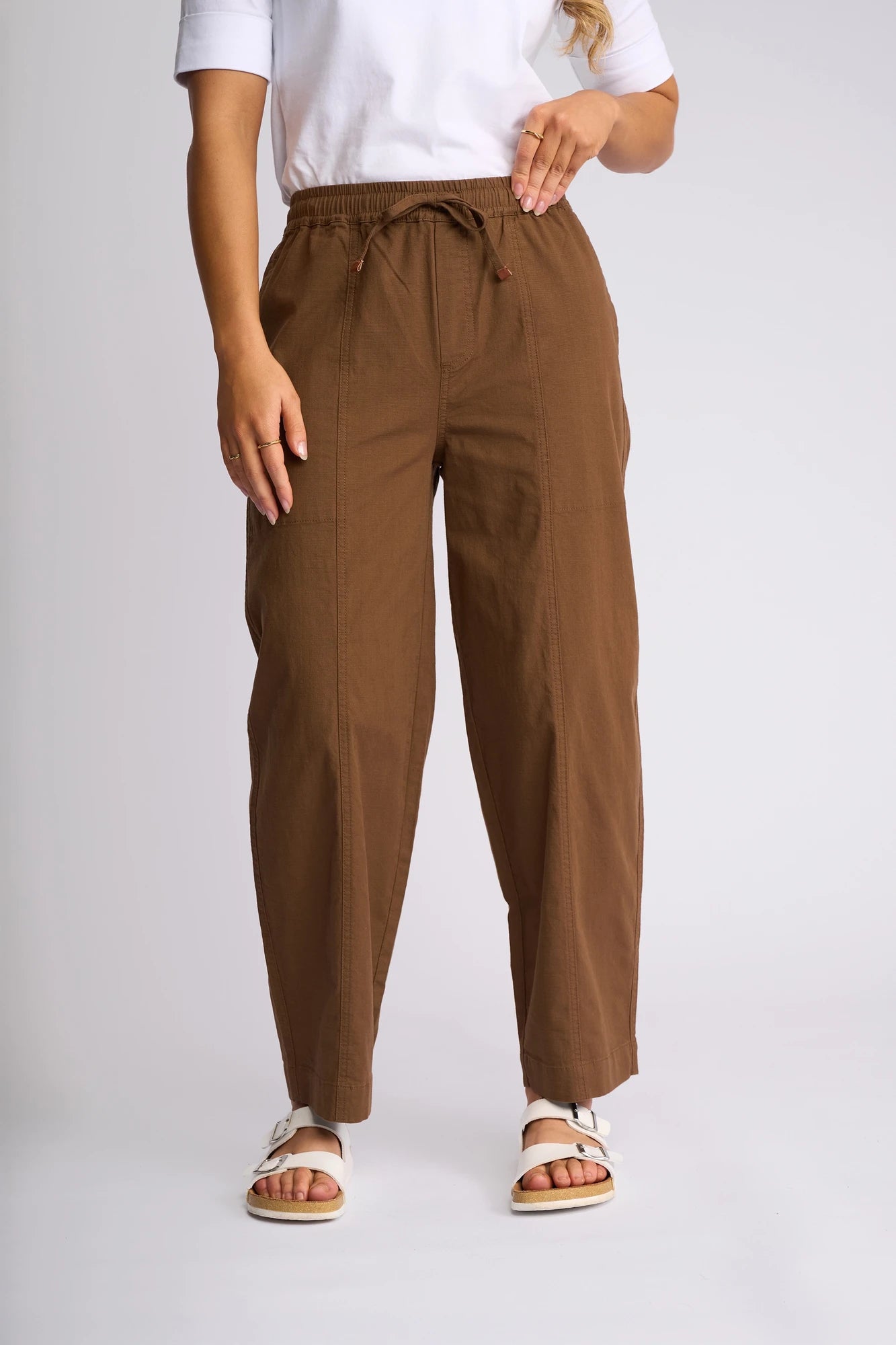 A person stands against a light gray background wearing a white shirt, white sandals, and the Vassalli Dione Pant in Pecan—featuring loose barrel legs and an elasticated drawstring waistband. The photo is cropped at the shoulders.